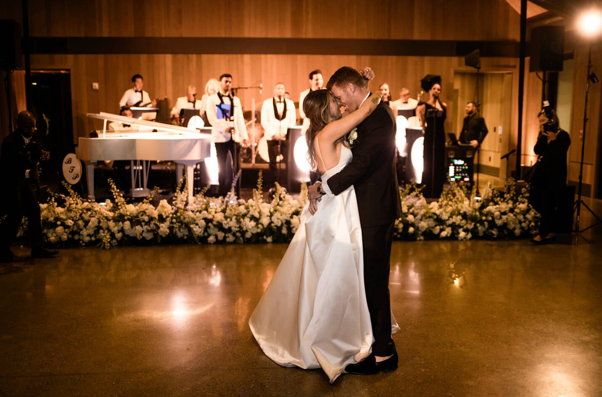 Bride and groom dance at wedding reception. Band plays on a stage decorated with flowers.