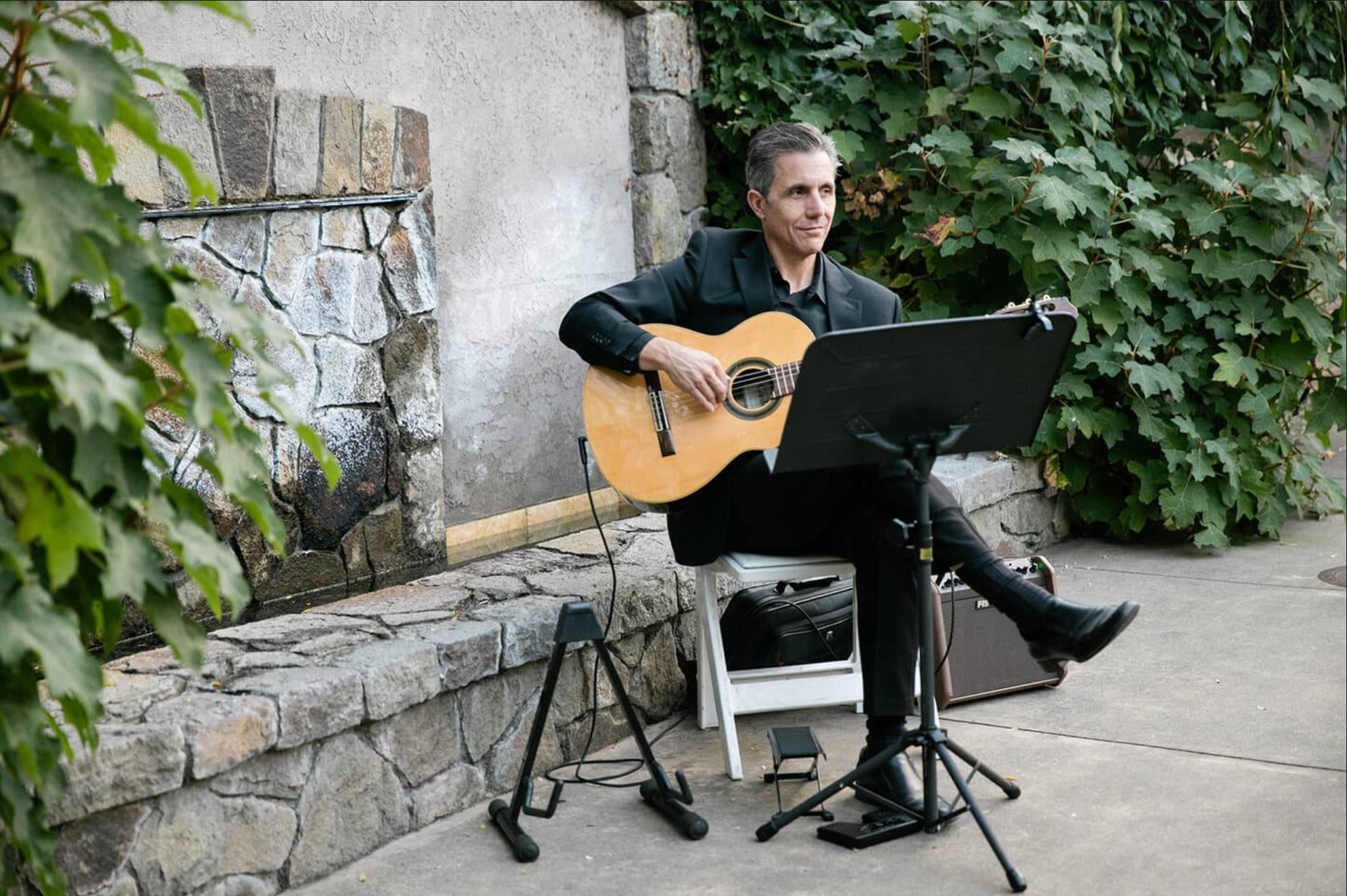Man playing acoustic guitar outdoors near a stone wall and foliage.