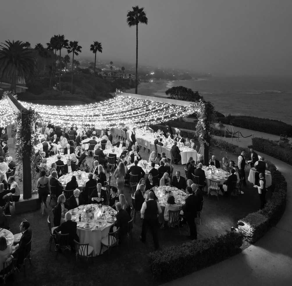 Outdoor wedding reception with guests seated at round tables under string lights, ocean view.