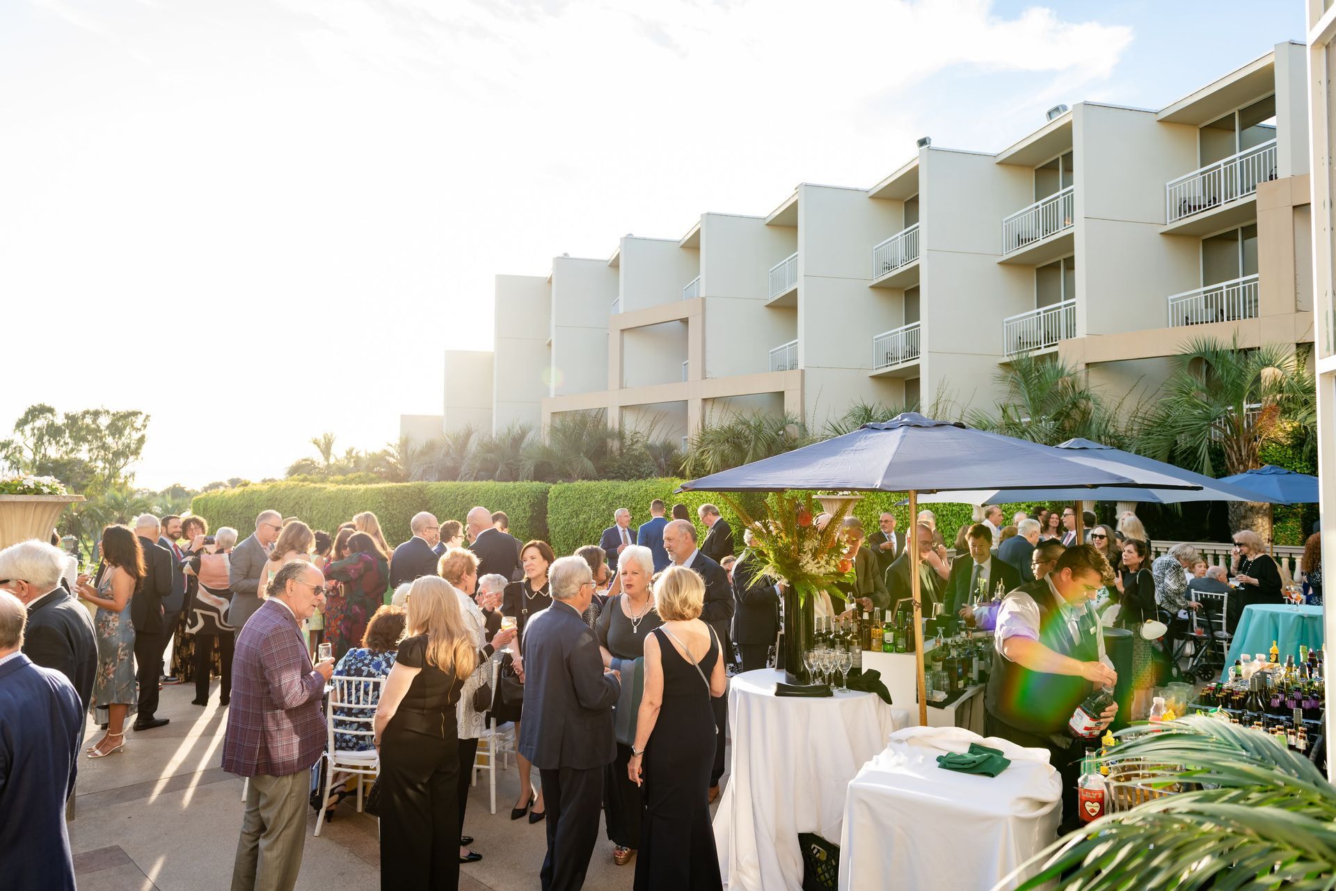 Outdoor evening event: crowd mingling near a hotel. People in formal attire, some near tables.