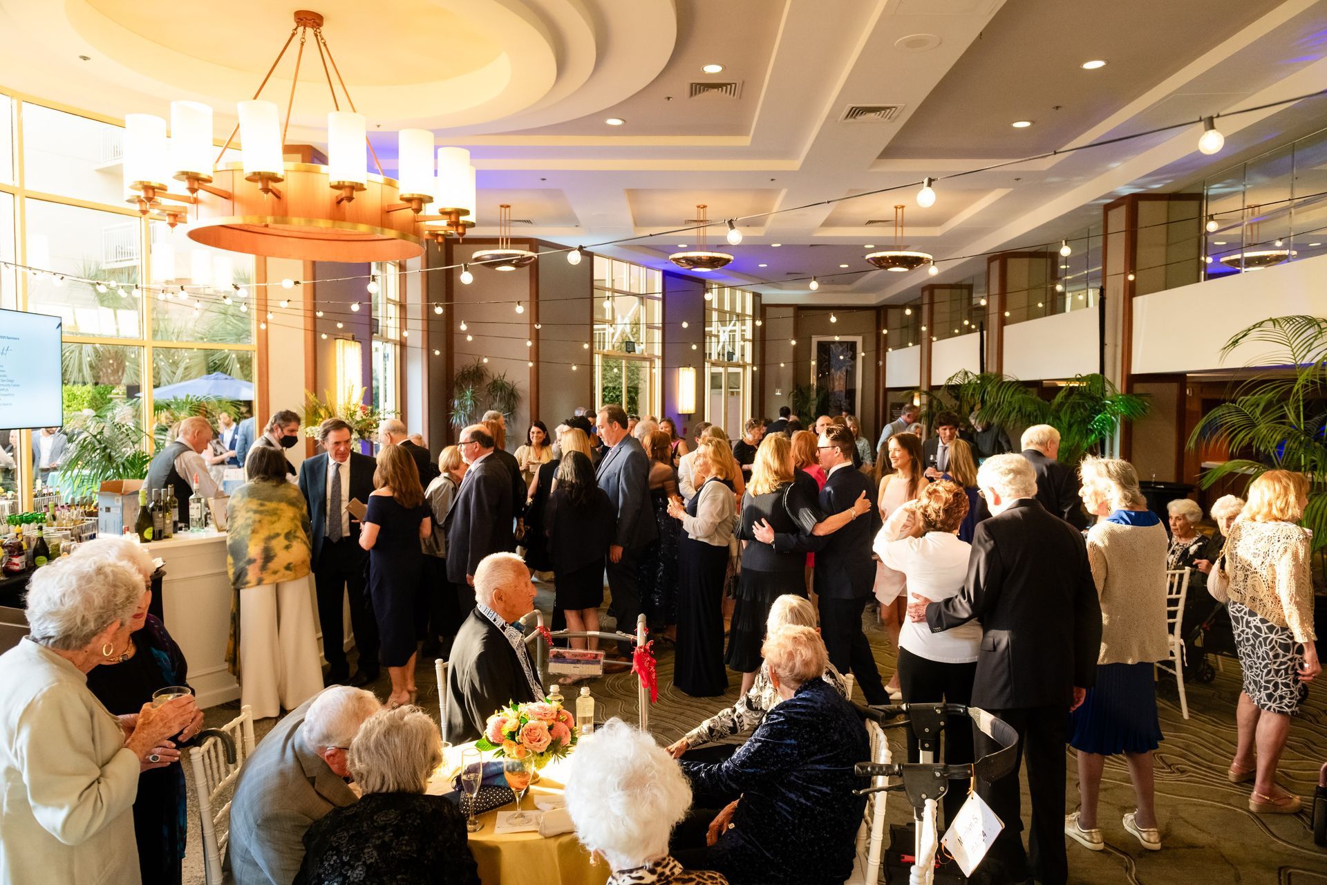 Guests mingle at a well-lit event in a ballroom with large windows.