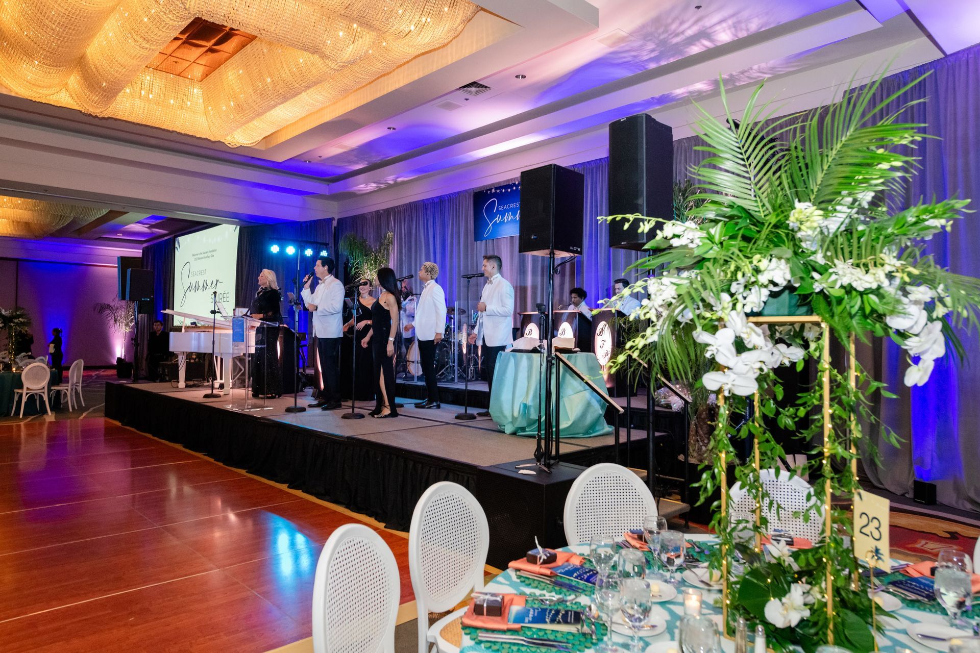 Band performing on stage at a gala. Elegant tables set with floral centerpieces and people in formal attire.