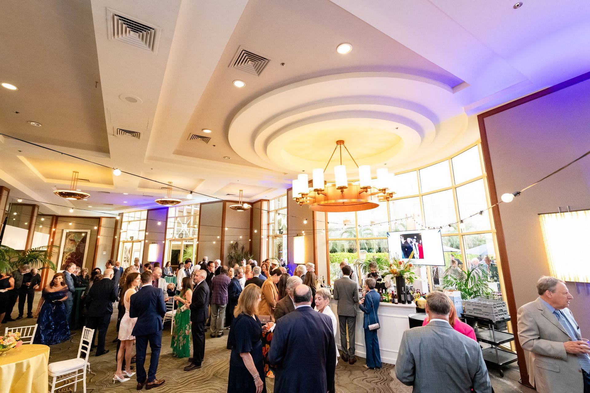 Large event room with a crowd. People in formal wear, a large chandelier, and outdoor view.