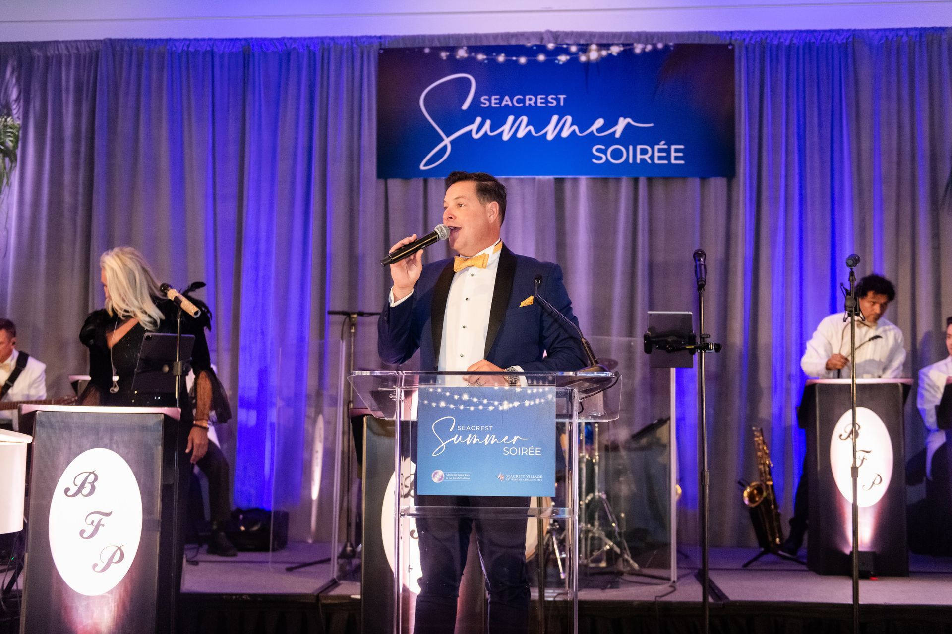 Band performs on stage at the Seacrest Summer Soirée. Singer in a tuxedo, blonde woman, and musicians with instruments.