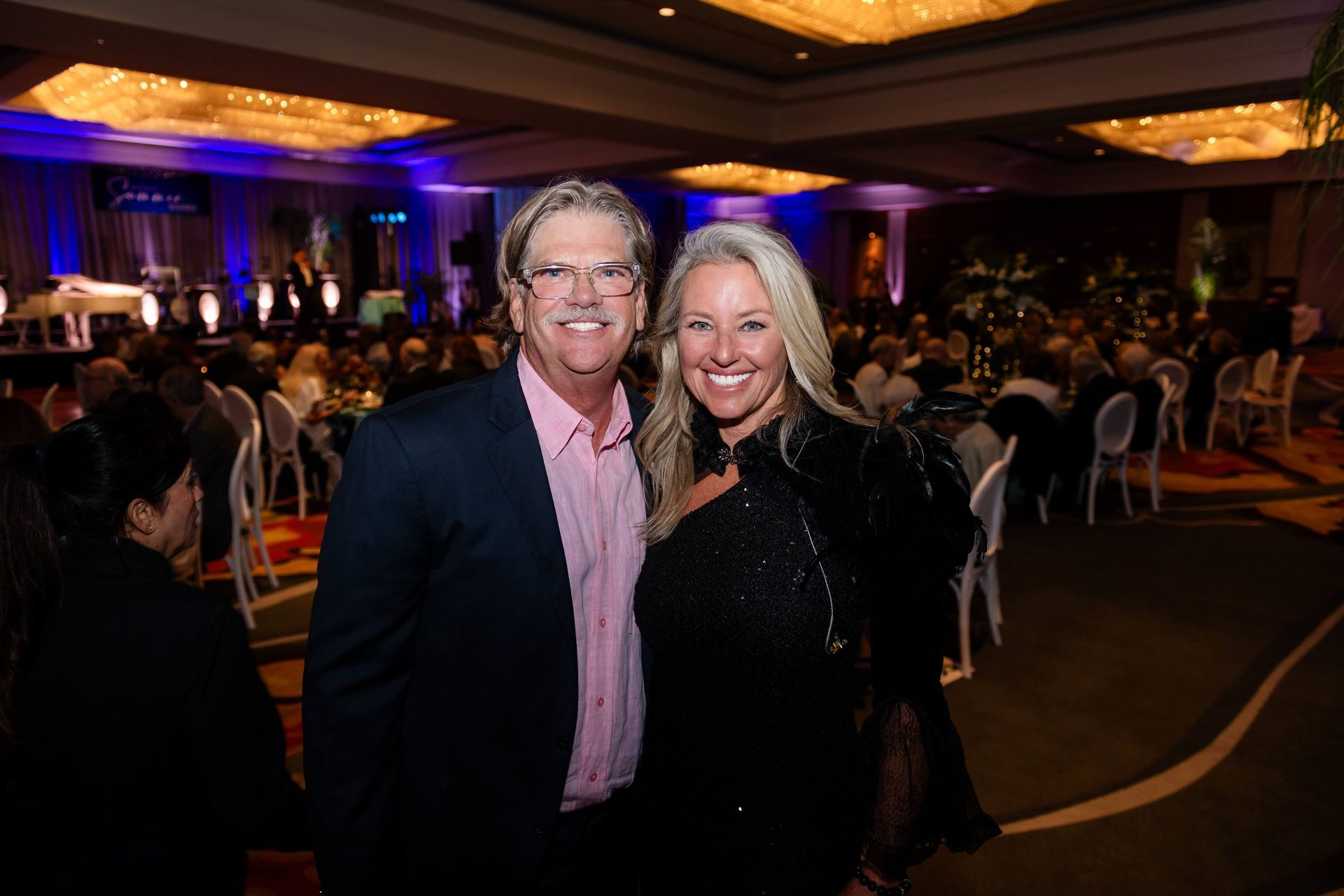 Man and woman smiling at a formal event; the man in a suit, the woman in a black dress, in a large ballroom.