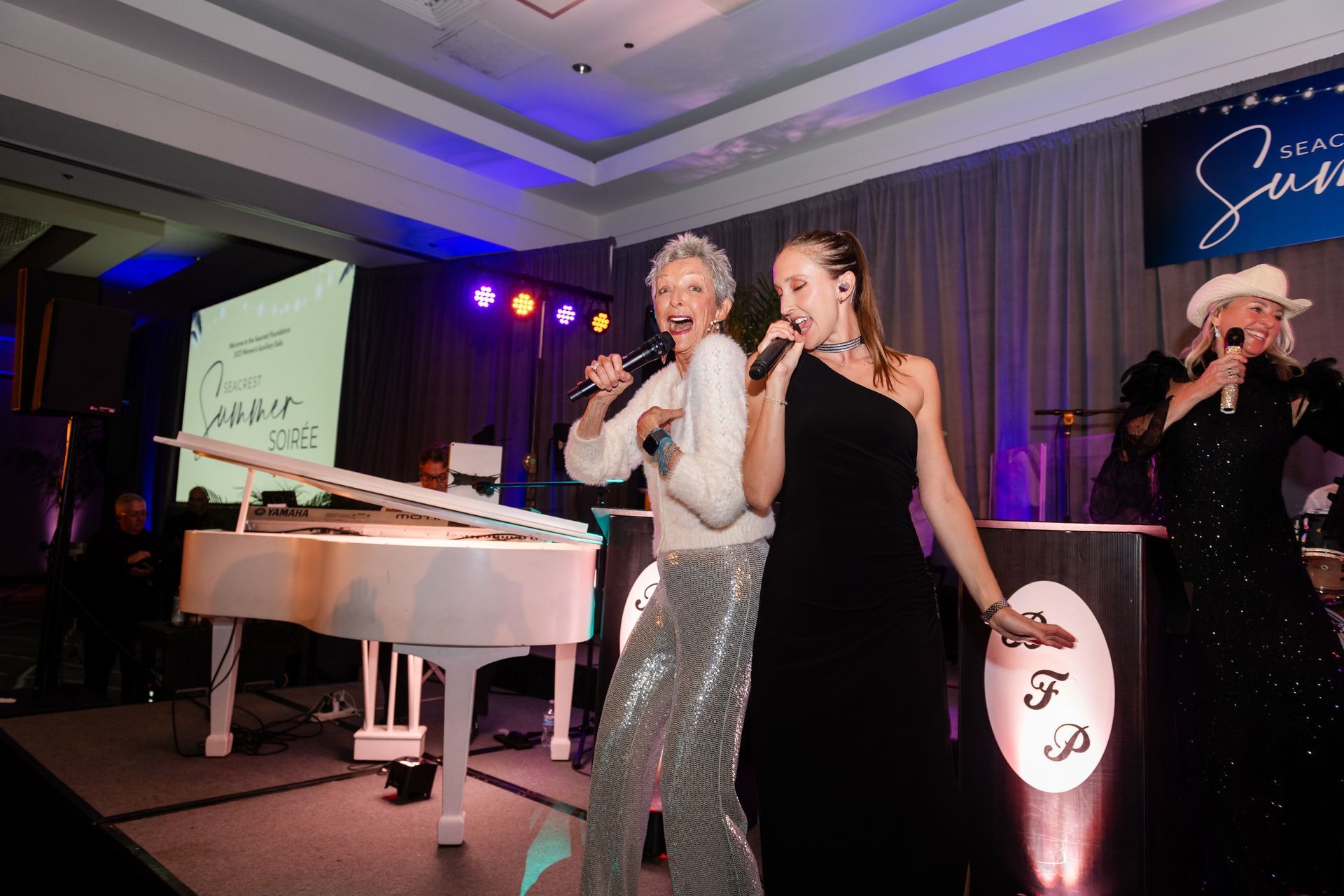 Three women singing onstage at an event, with a white piano and a backdrop.