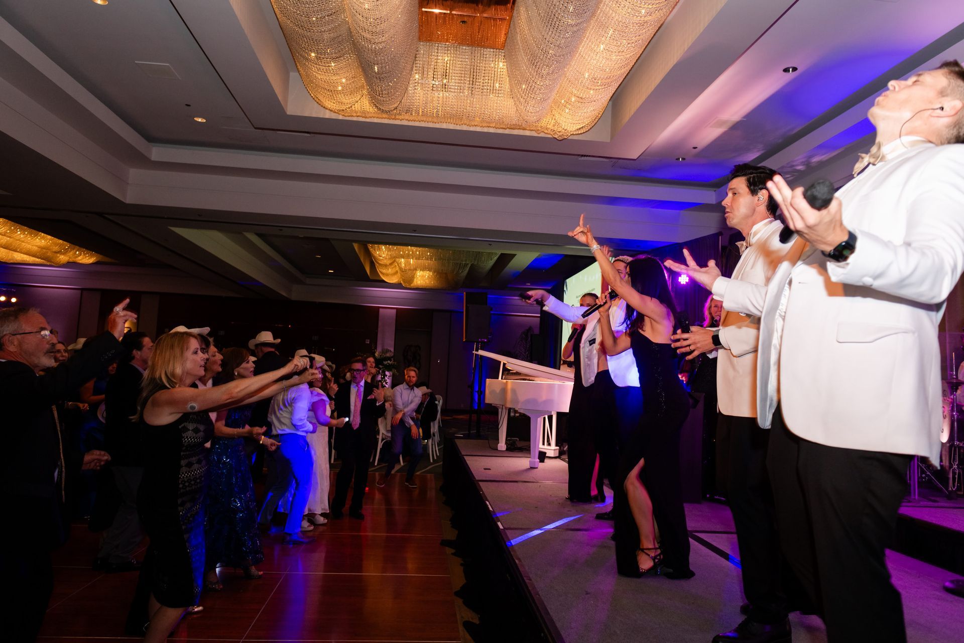 People dancing and singing on stage at an event, with a band, colorful lights, and a chandelier.