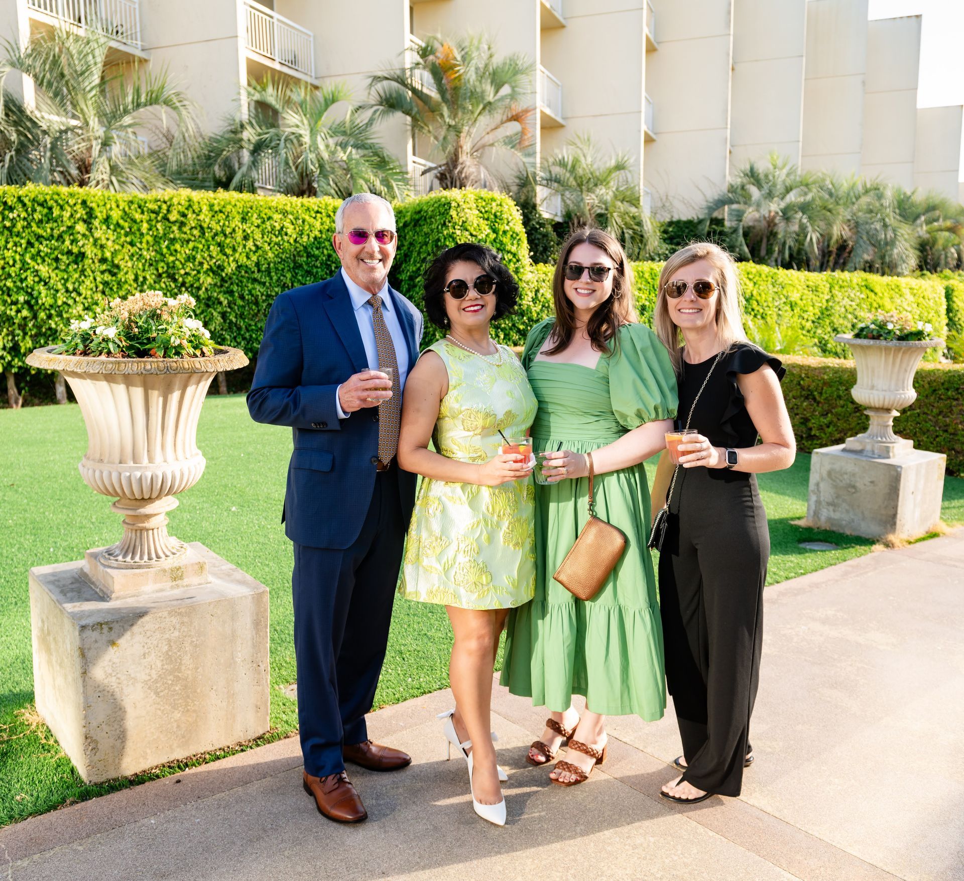 Four people smiling and posing outdoors with drinks; a man, two women, and another woman.