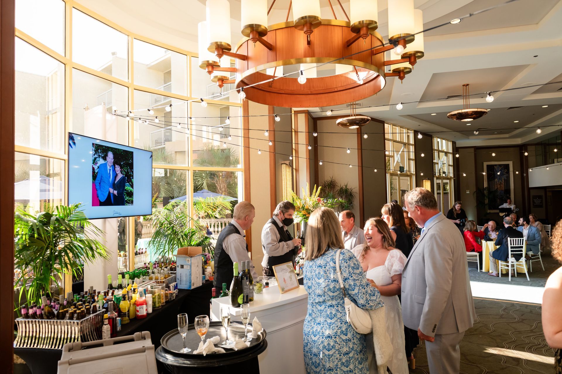 Elegant event with bar, people mingling, large windows, and a chandelier.