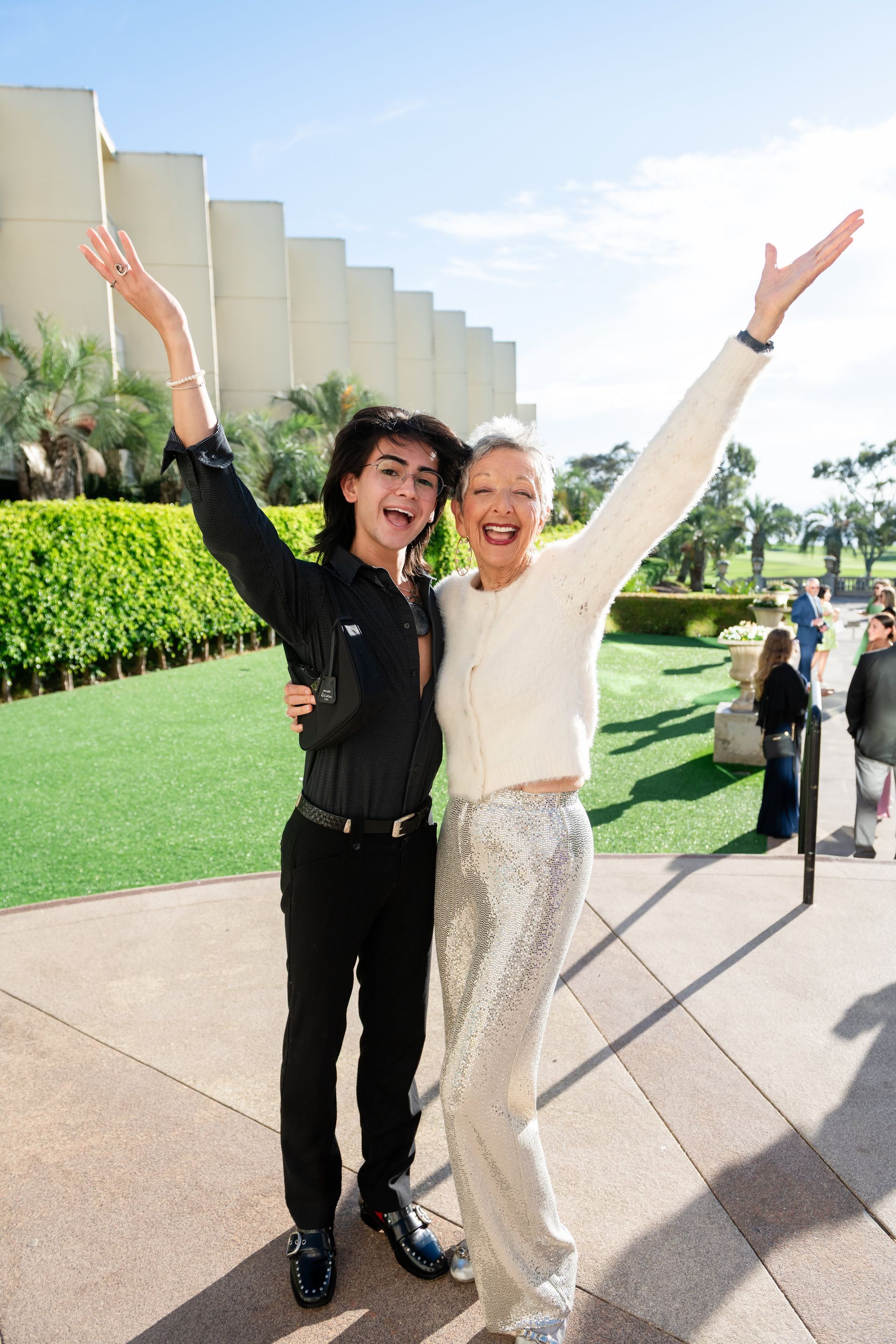 Two people smiling with arms raised outside; one in black, one in white sequins, sunny.