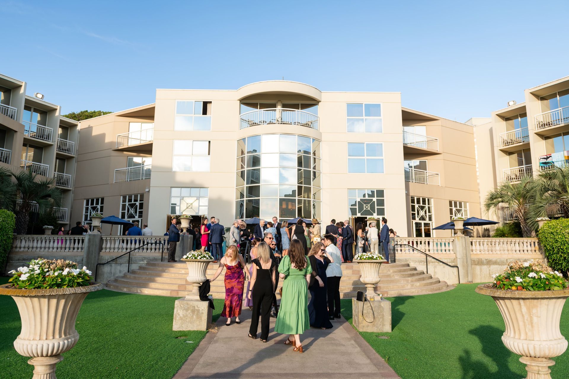 Guests in formal attire gather outside a beige hotel with large windows; flower pots line the lawn.
