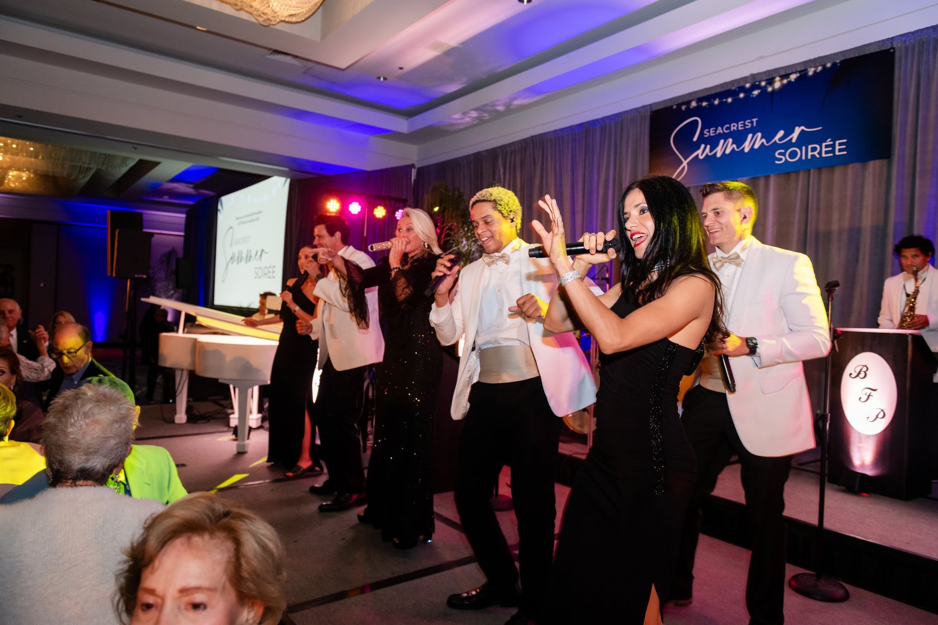 Band performing on stage at a summer soiree, with formal attire and microphones, people in audience.