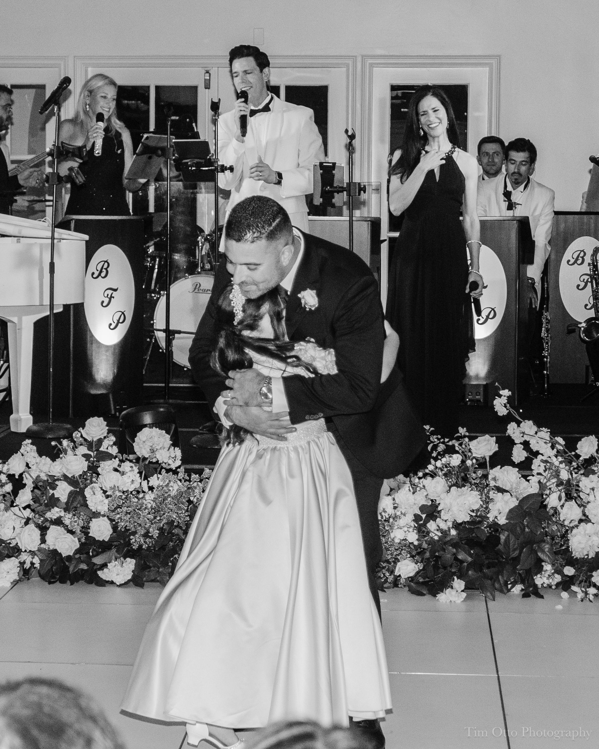 Groom hugs flower girl on a dance floor, a band performing in the background. The setting appears to be an indoor wedding reception.