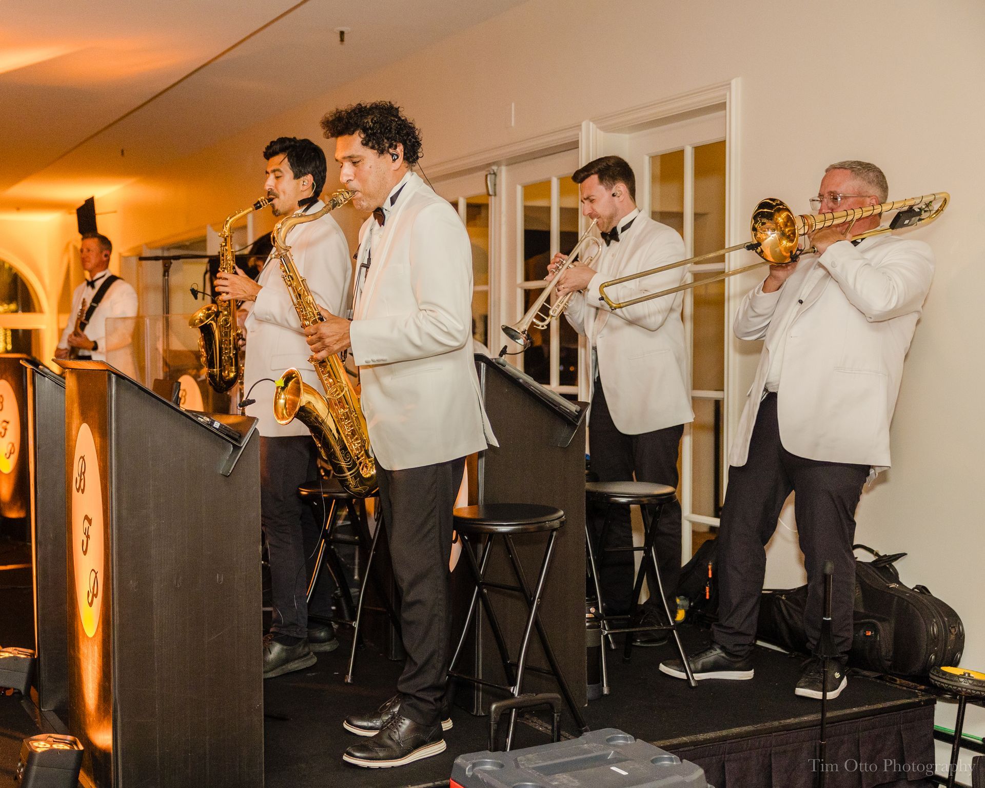 A live band horn section of six men in white jackets performs on a stage, featuring saxophones, trombone, and trumpet. They are playing at an indoor event.