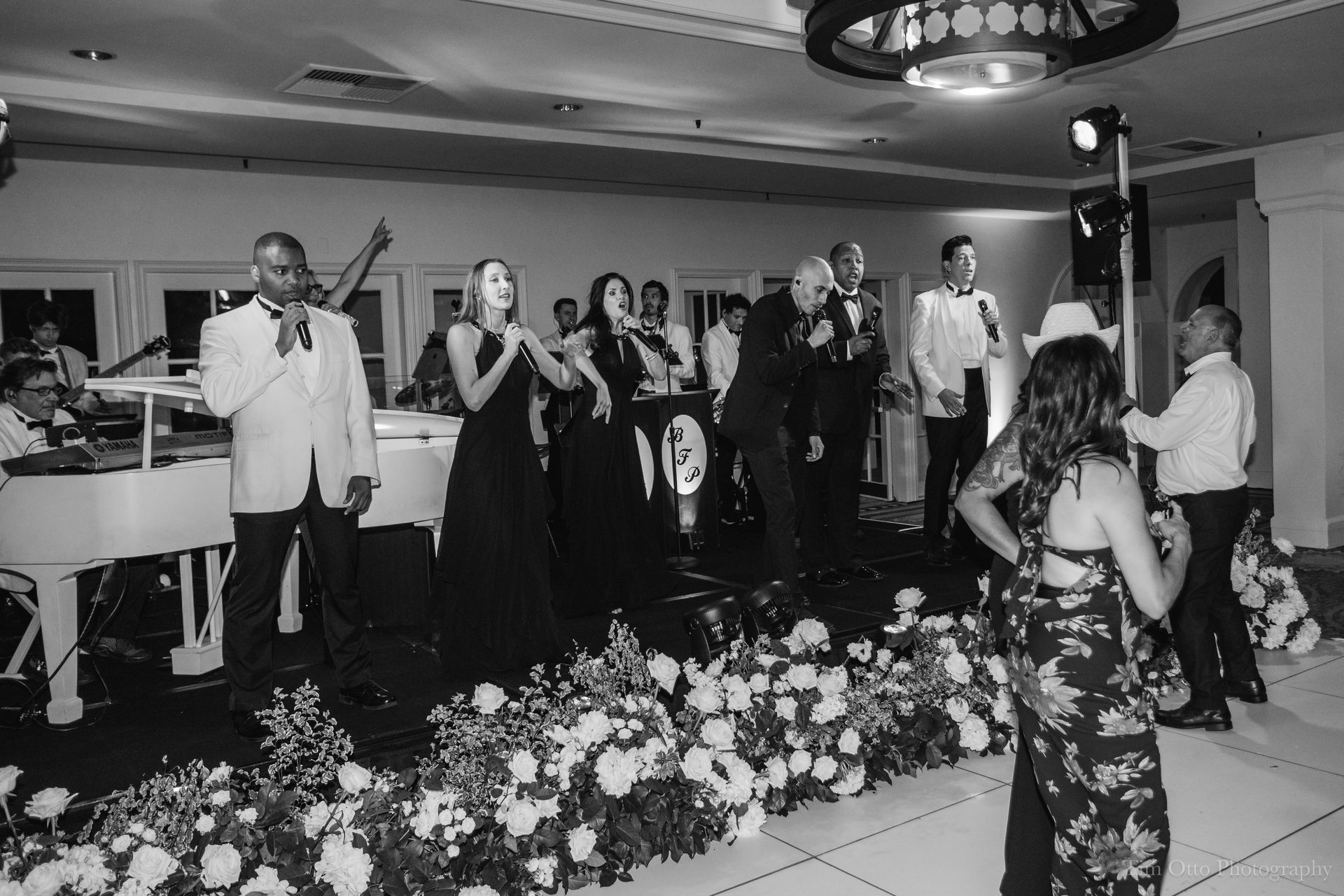 A band performs on a stage at a wedding. Several singers with microphones are performing in front of a white piano, and some guests are dancing.
