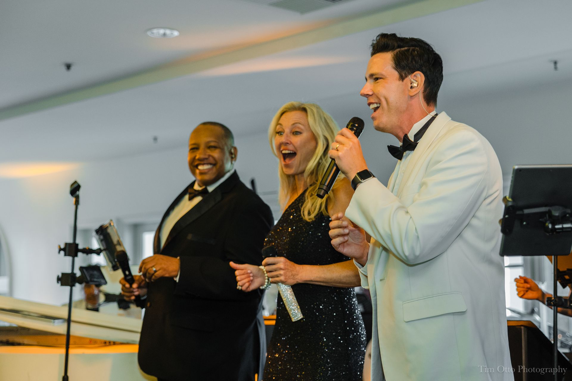 Three singers on a stage: a Black man, a blond woman, and a white man, all smiling and singing. The man on the right is holding a microphone.