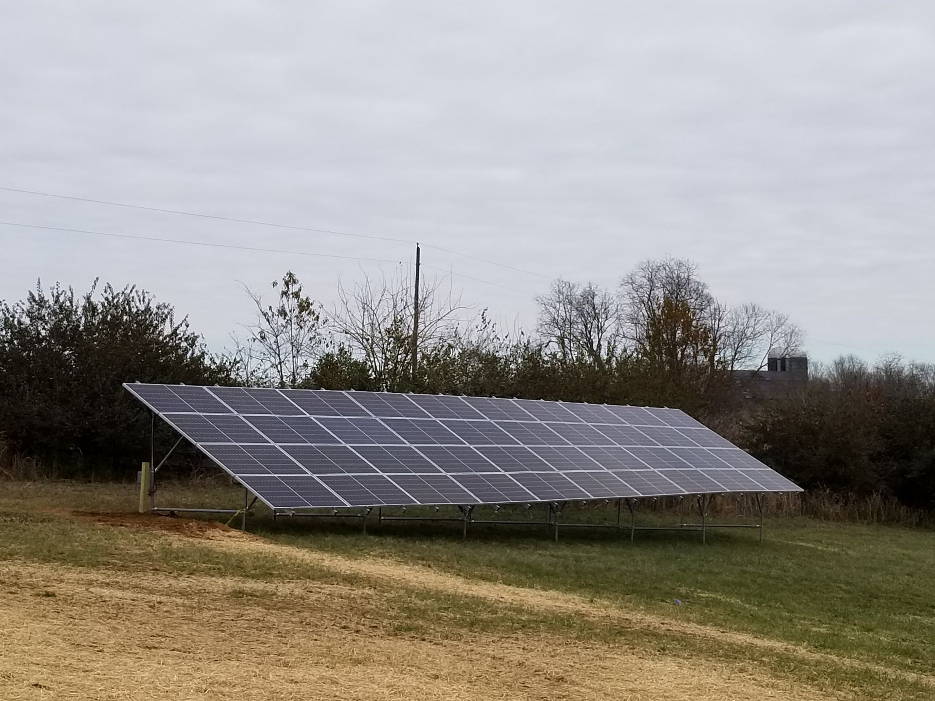 A ground-mounted array of solar panels positioned in a grassy field under a cloudy, overcast sky.