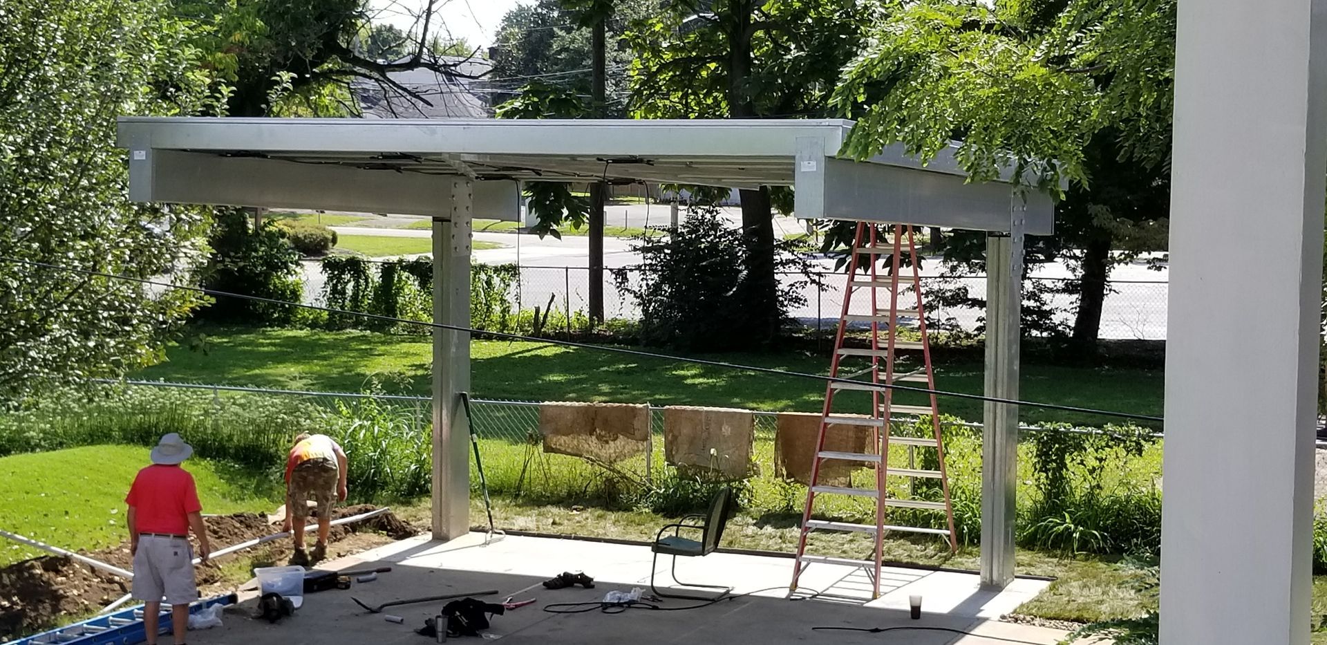 Workers building a carport outdoors; ladder and chairs nearby.