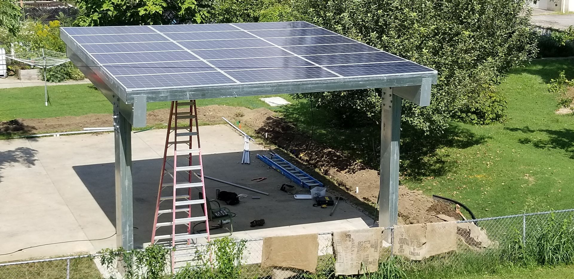 Solar panel canopy over a concrete pad, with a ladder and tools present. Green grass and trees are in the background.
