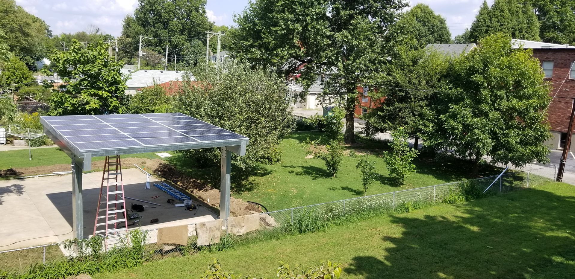 Solar panel carport in a grassy yard, surrounded by trees and a building. Ladder propped up inside.