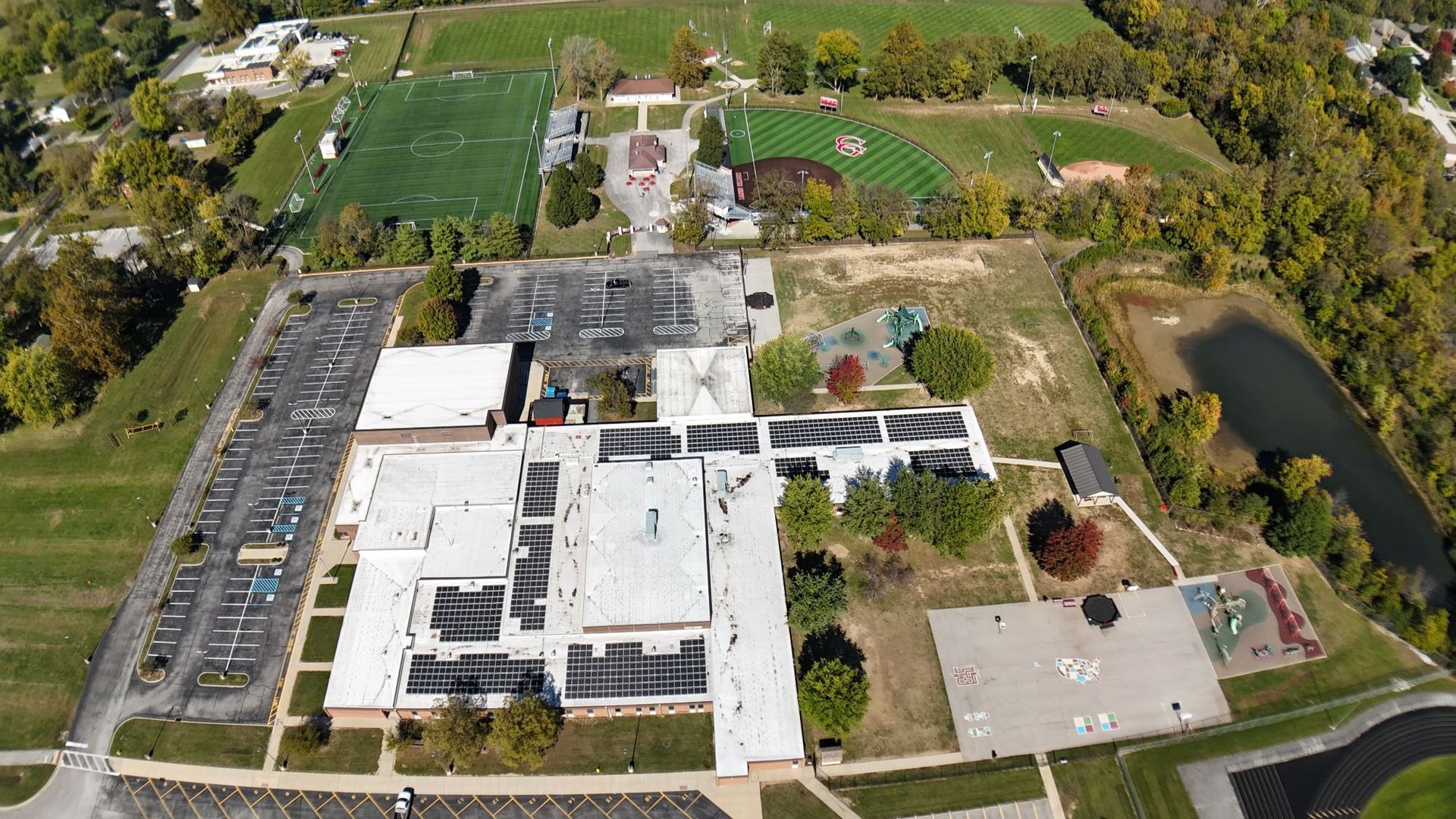 Aerial view of school building with parking lot, sports fields, and pond.