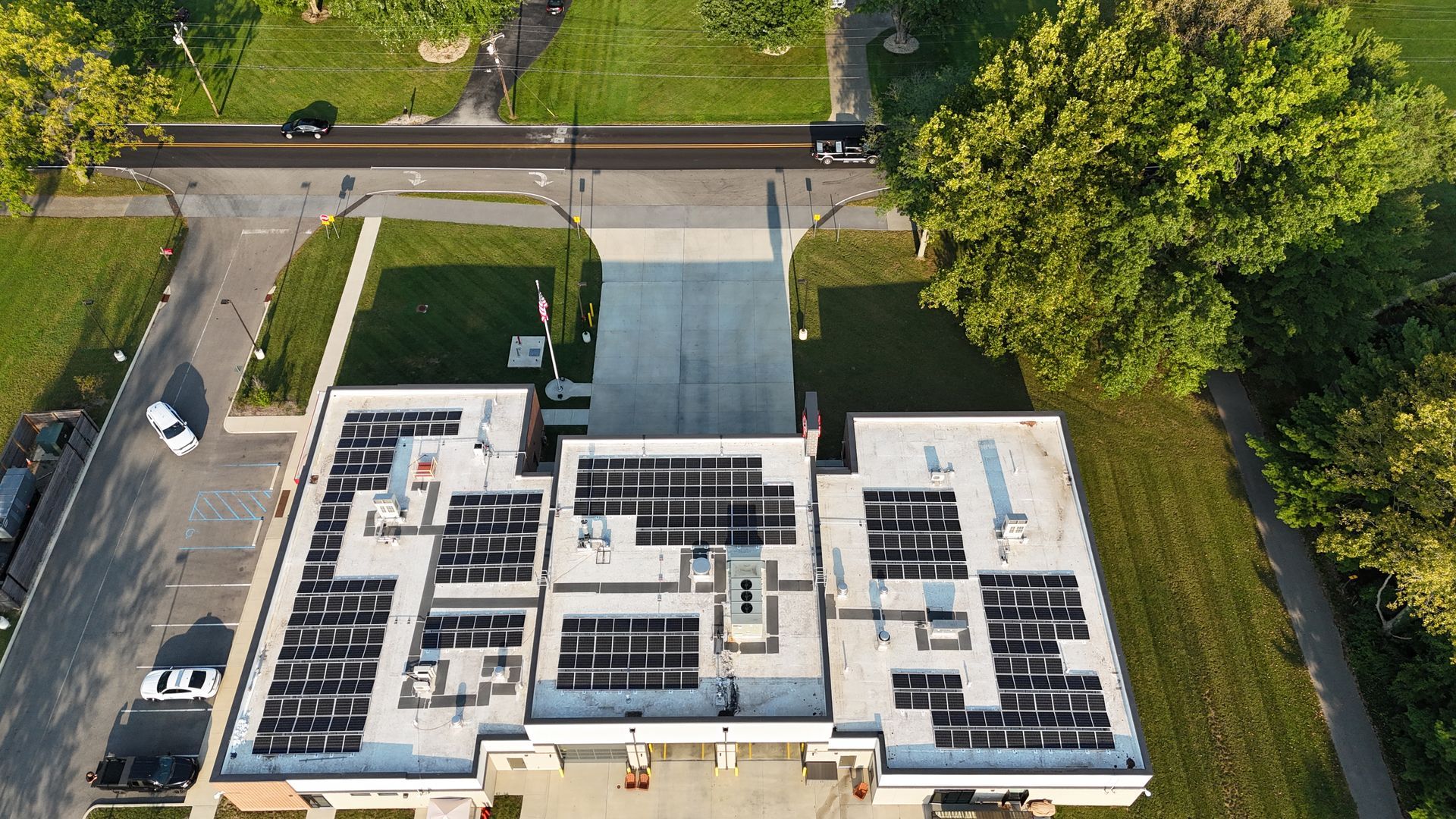 Aerial view of building with solar panels on the roof, driveway, road, and trees.
