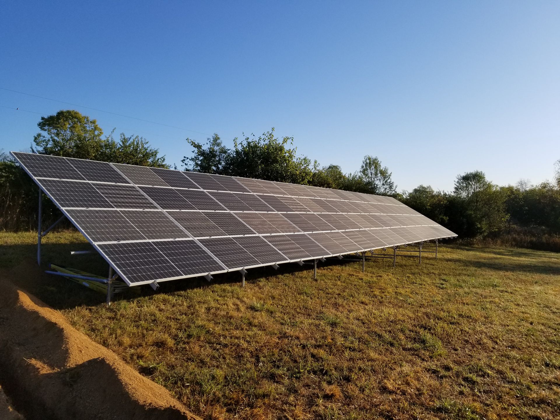 Solar panel array on a metal frame in a grassy field, trees in the background, blue sky overhead.