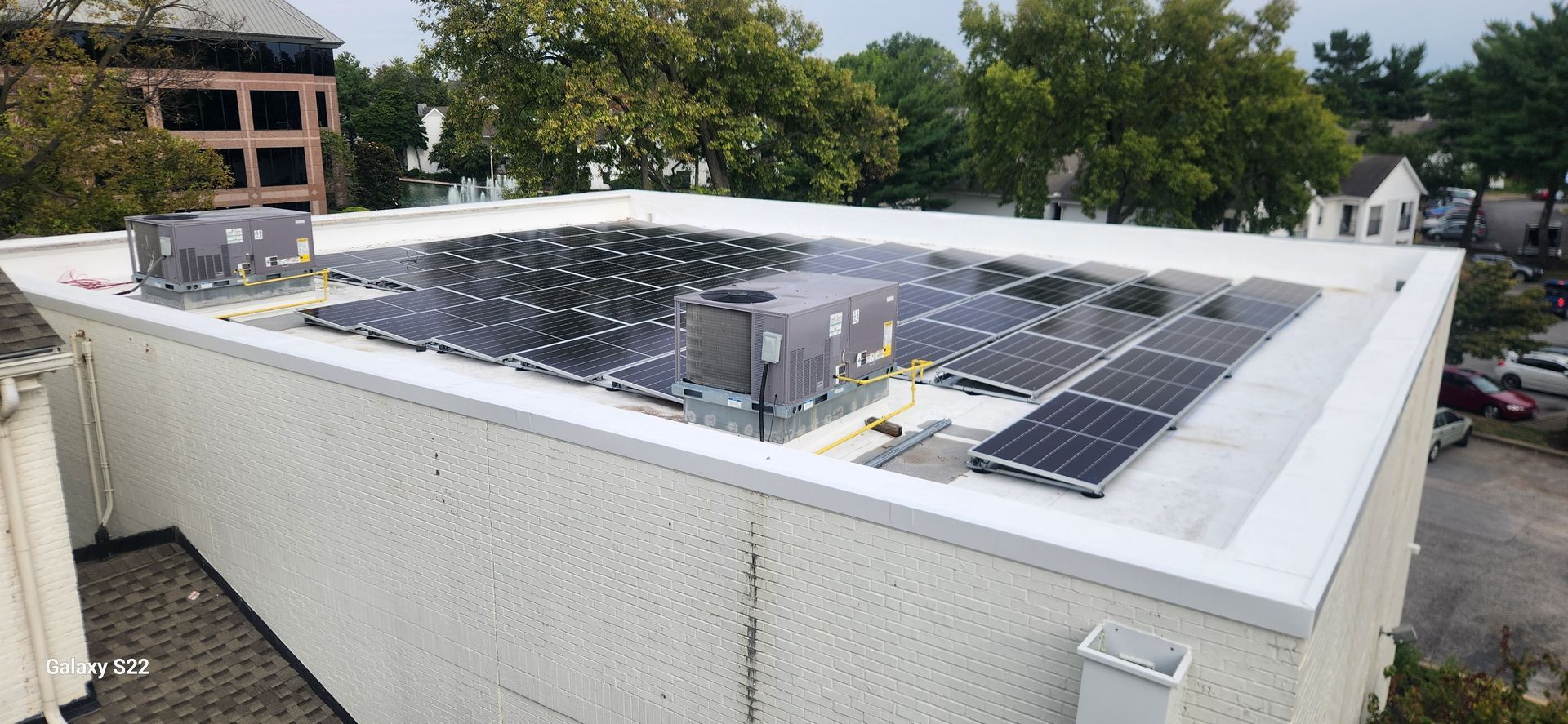 Solar panels on a white flat roof of a commercial building. Lush green trees in the background. Cloudy day.