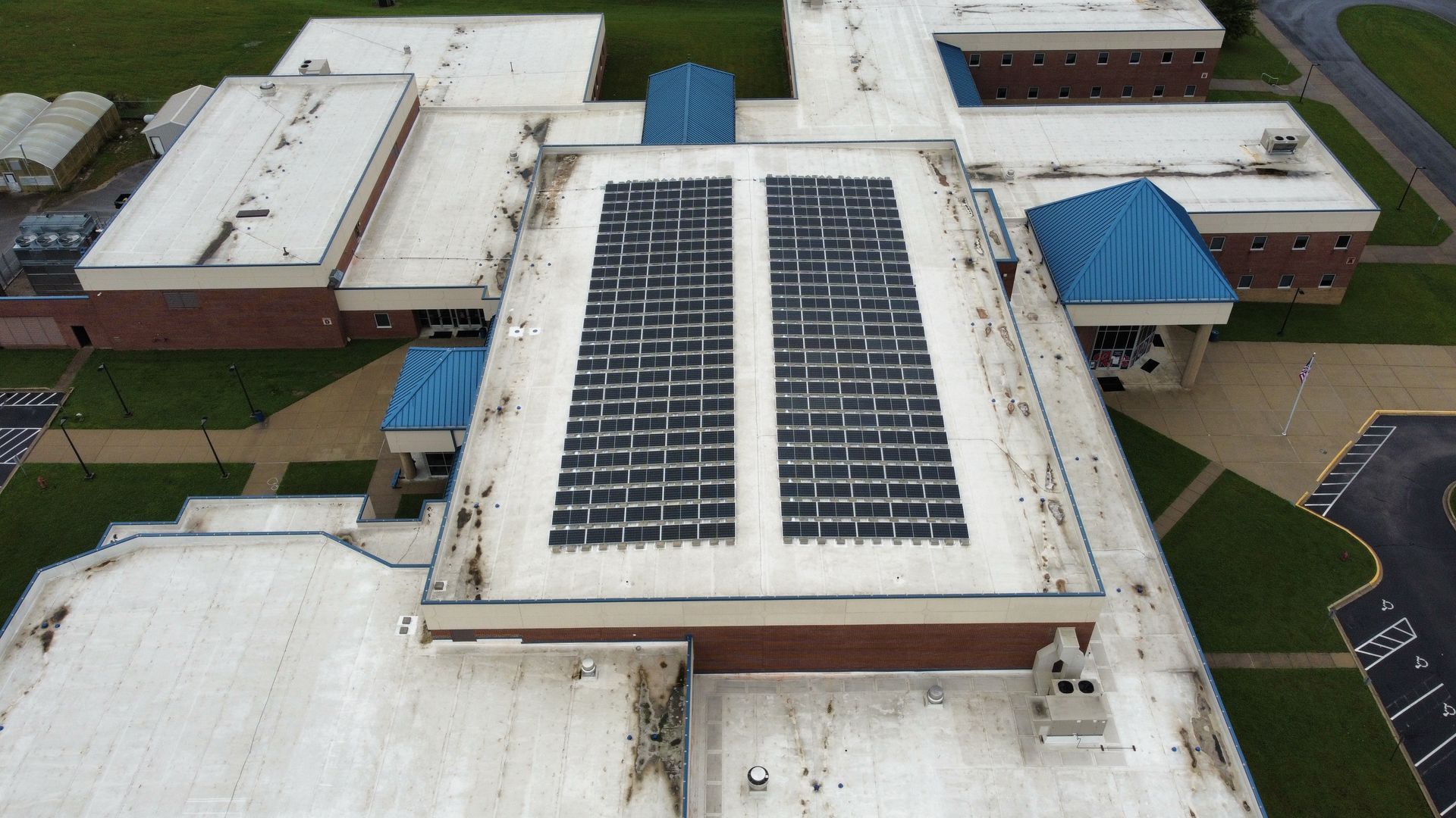 Overhead view of a school roof with solar panels installed. The roof is white with some blue coverings and surrounded by green grass.