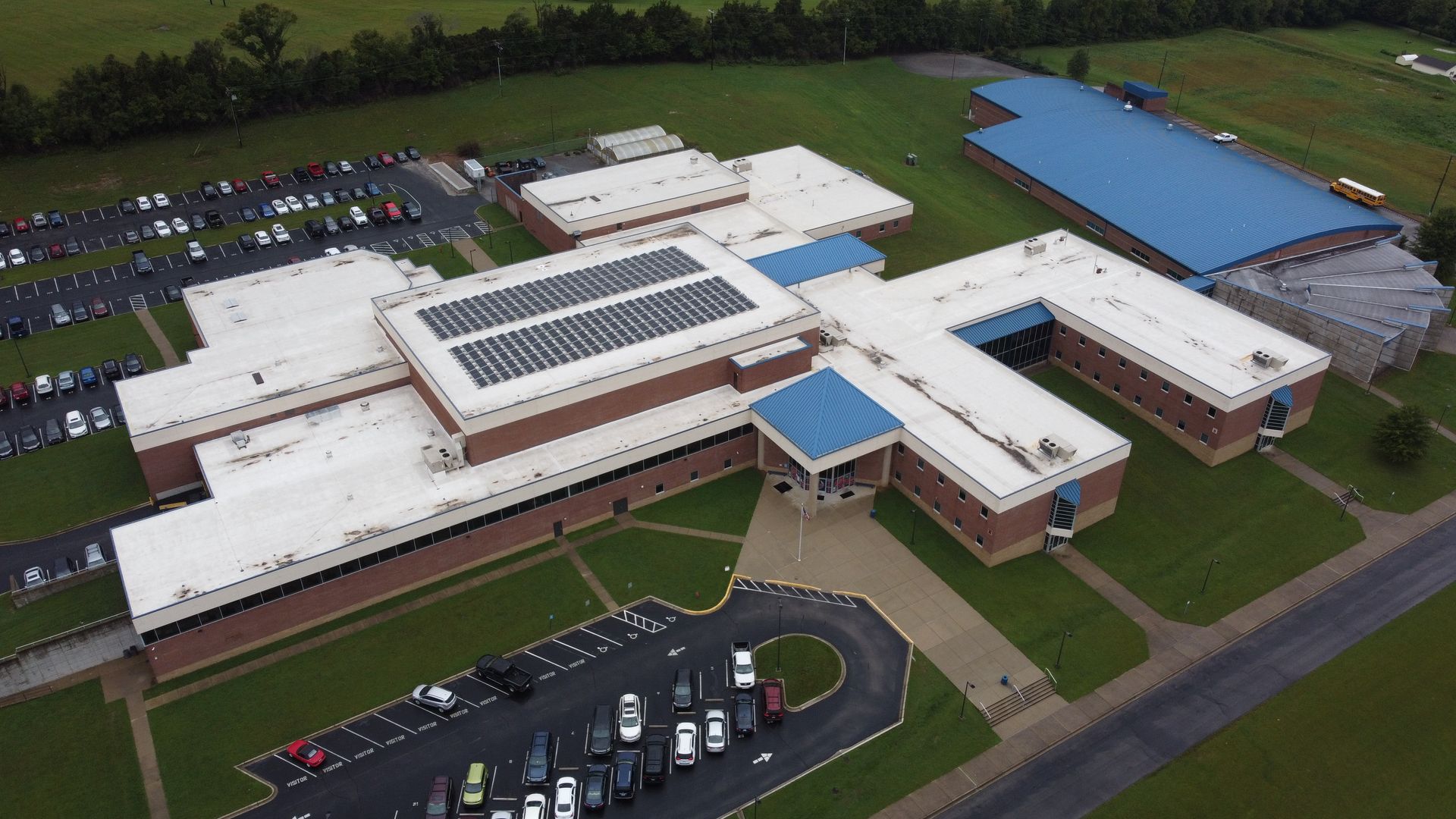 Aerial view of a school building with solar panels on the roof, surrounded by parking lots and green fields.