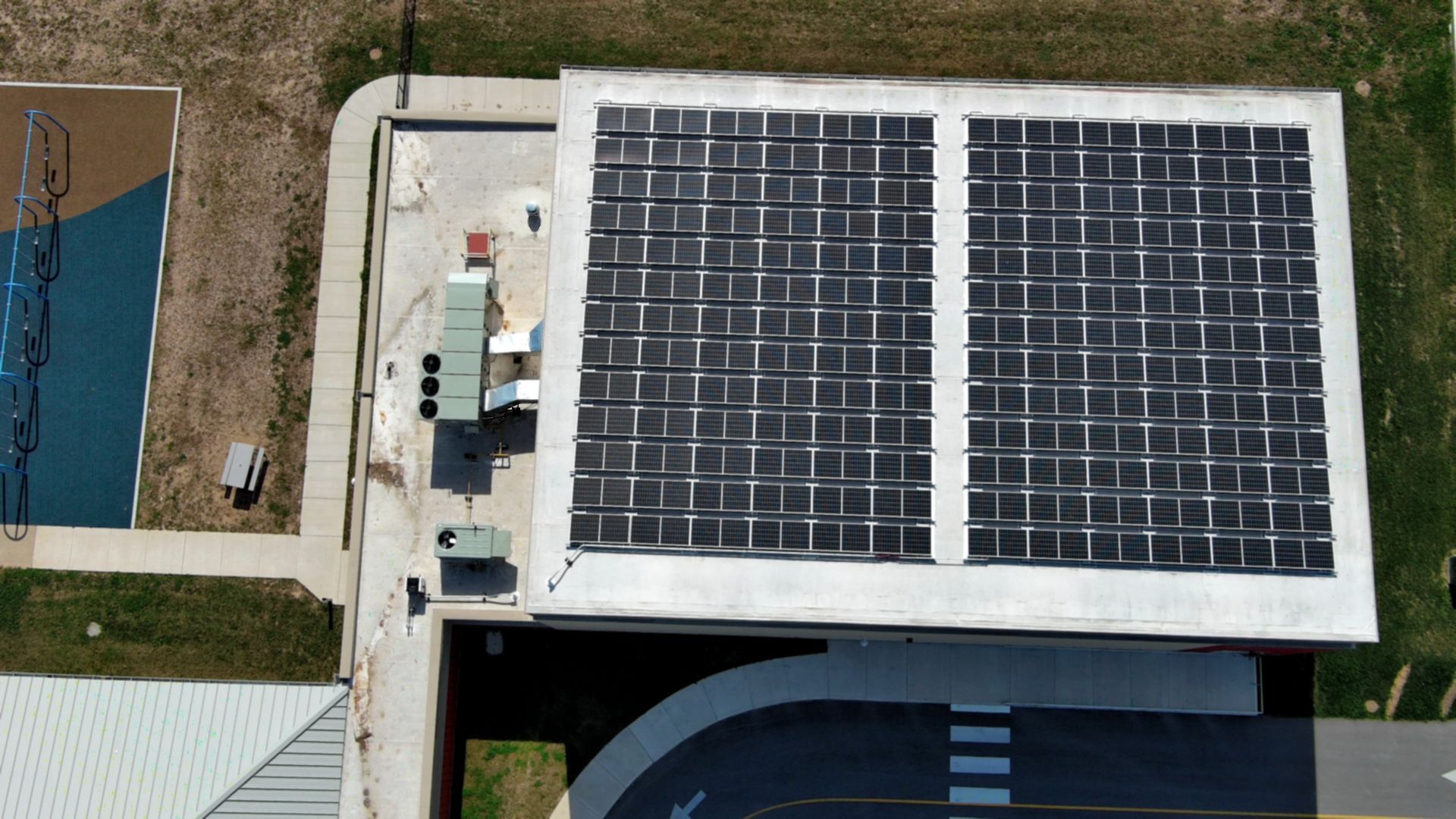 Overhead view of a building roof covered with solar panels, next to a playground and road.
