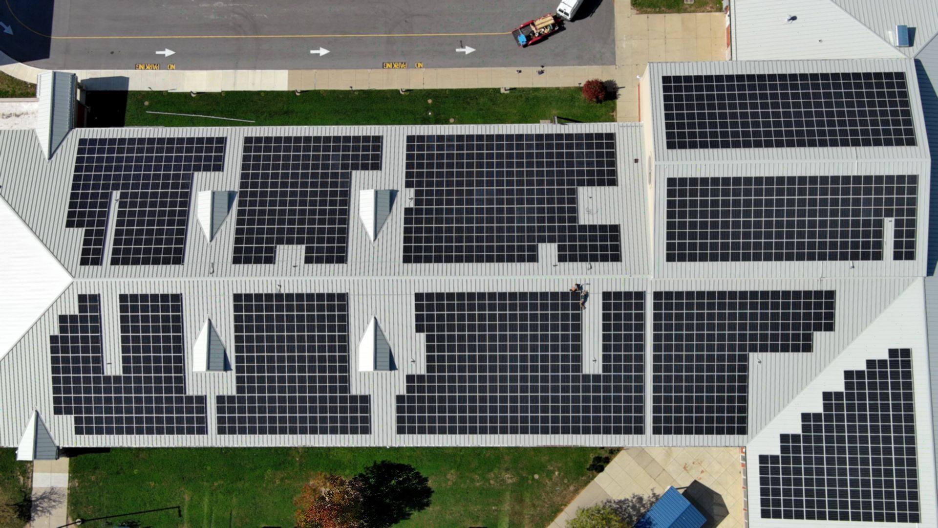 Aerial view of a building roof covered with numerous solar panels, near a road, grass, and other buildings.
