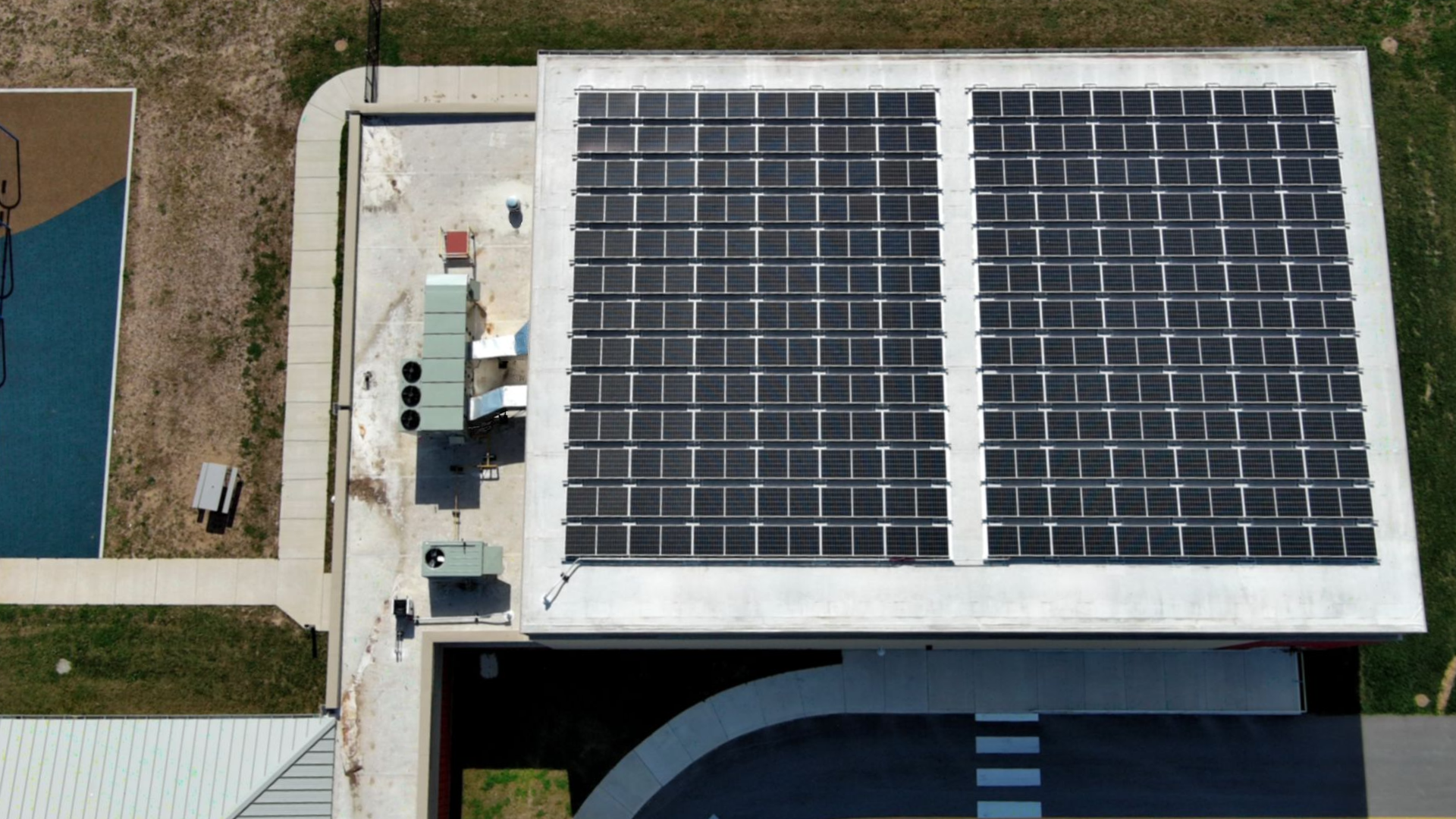 Overhead view of a building roof covered with solar panels, next to a playground and road.
