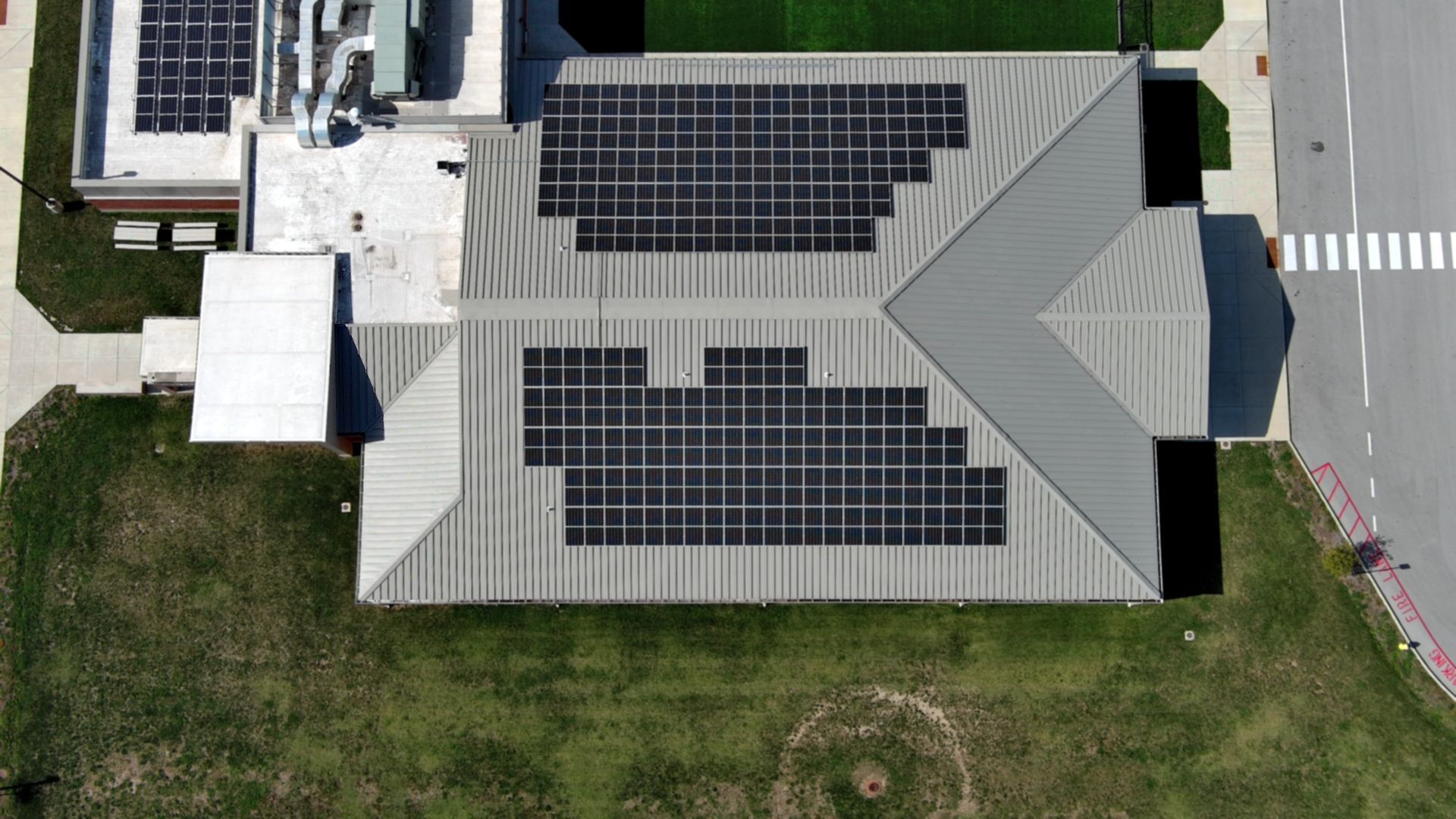Overhead view of a building roof with multiple solar panel arrays. Gray roof, green lawn, crosswalk.