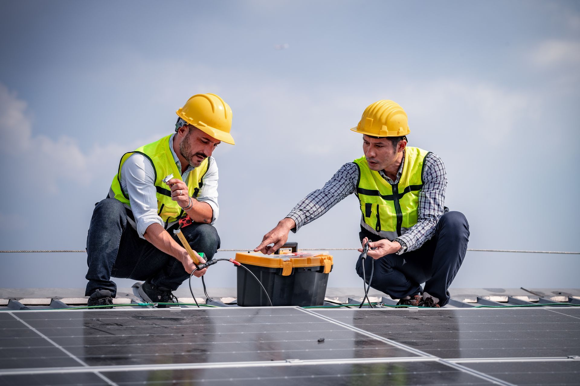 Two solar panel installers on a roof, connecting wires near a toolbox. They wear yellow hard hats and vests.