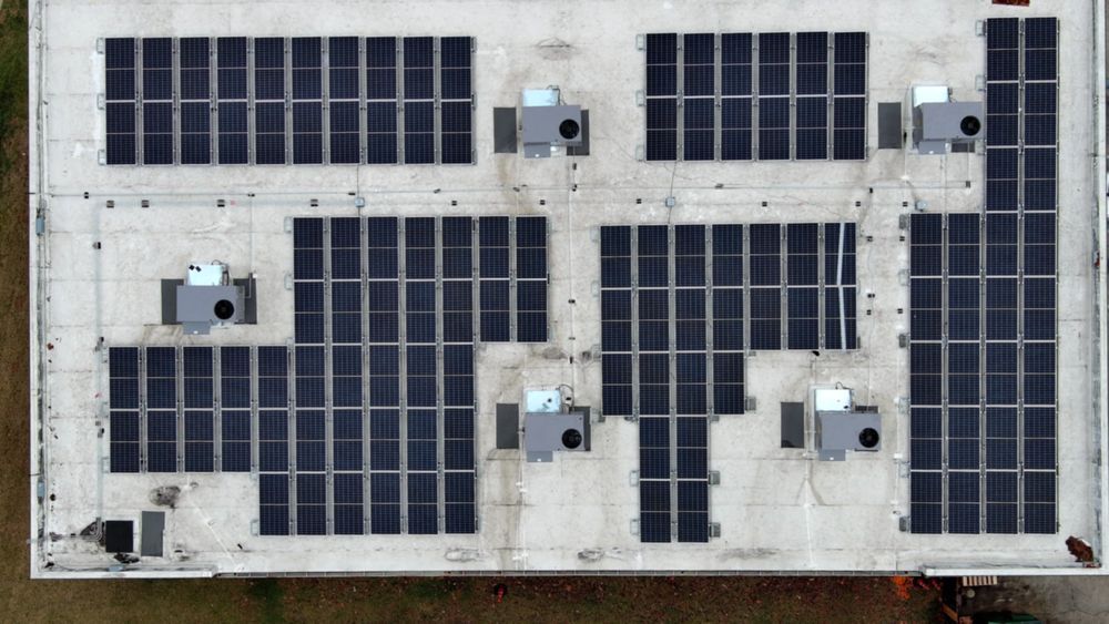 Aerial view of a building rooftop with solar panels and HVAC units installed. Gray and black colors dominate.