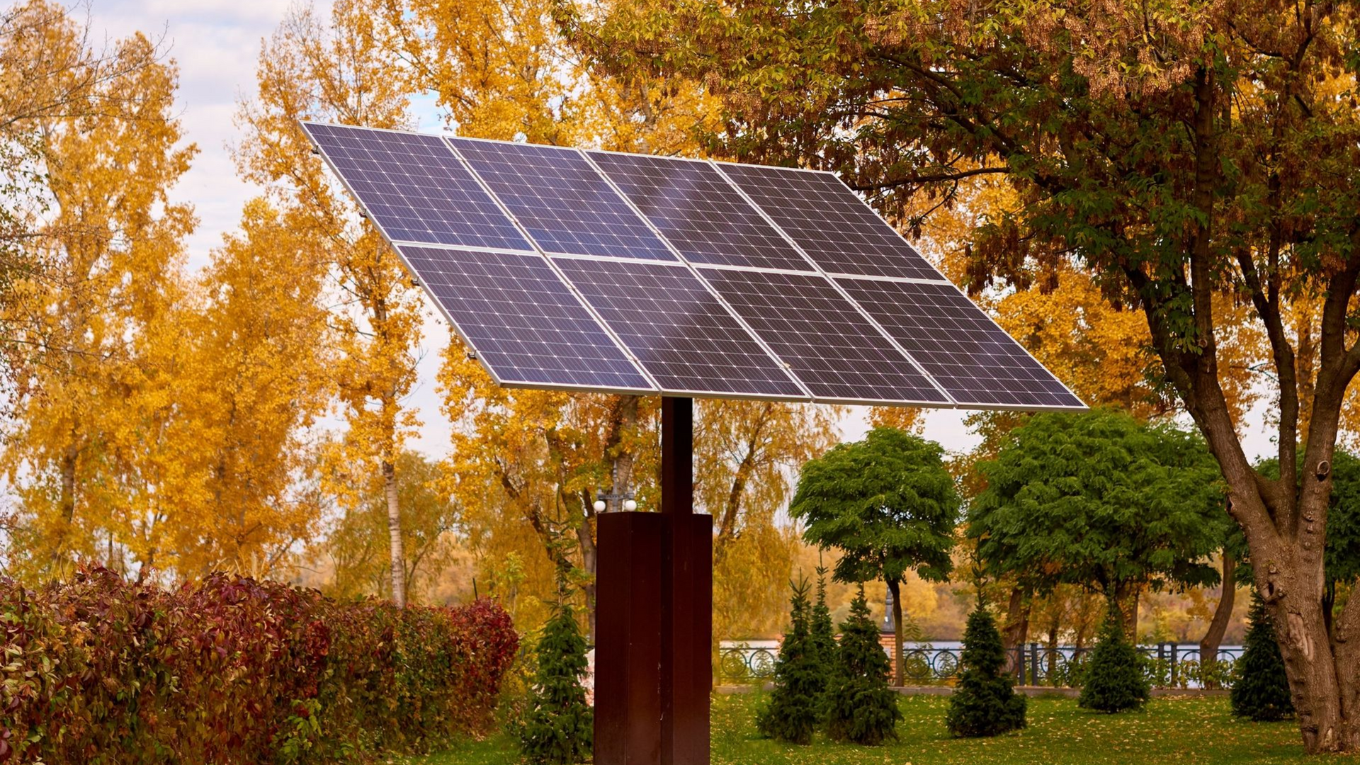 Solar panel in a park, surrounded by yellow autumn trees and green grass under a cloudy sky.