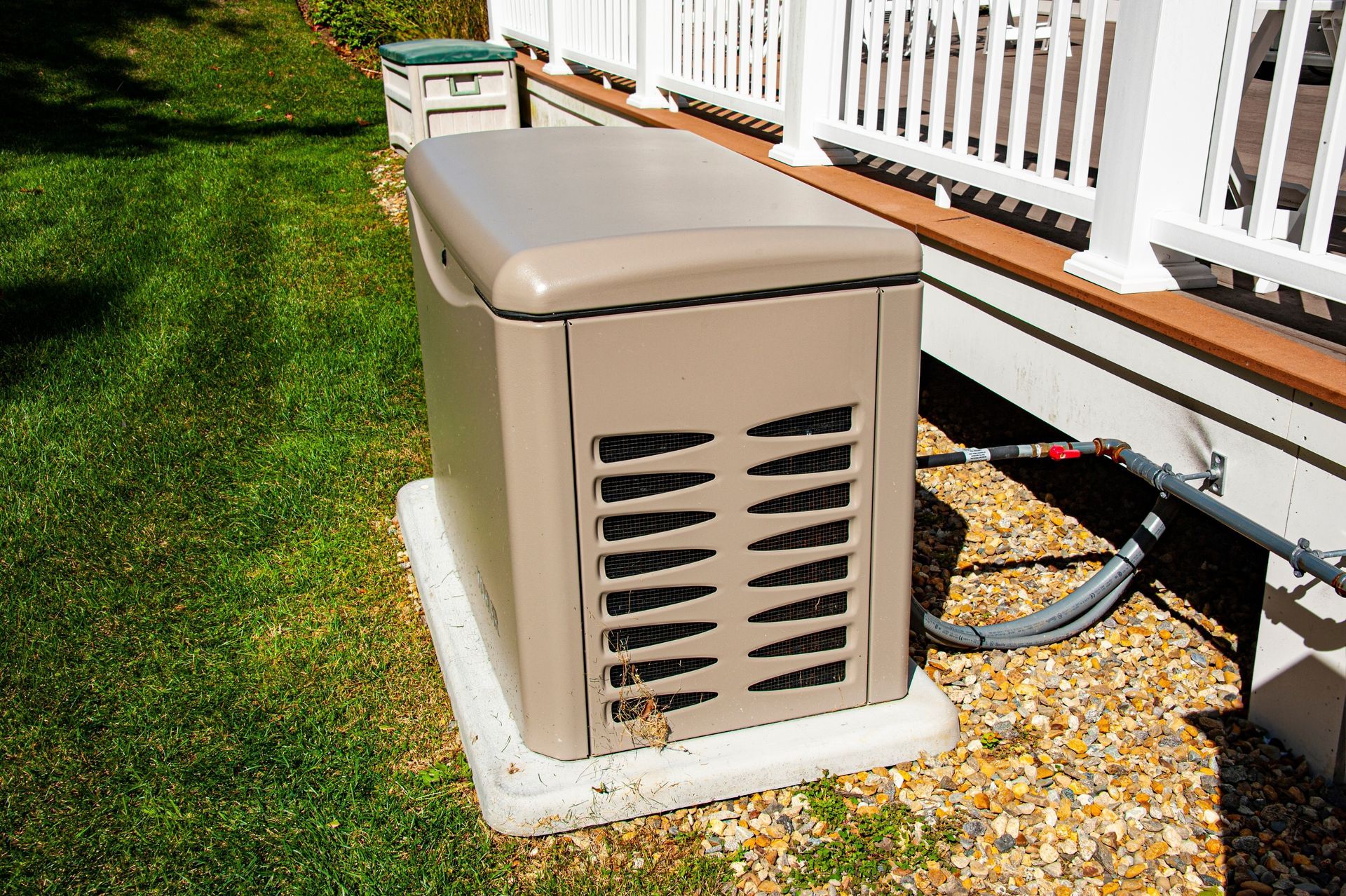 Beige home generator on a concrete pad next to a white deck on green grass.