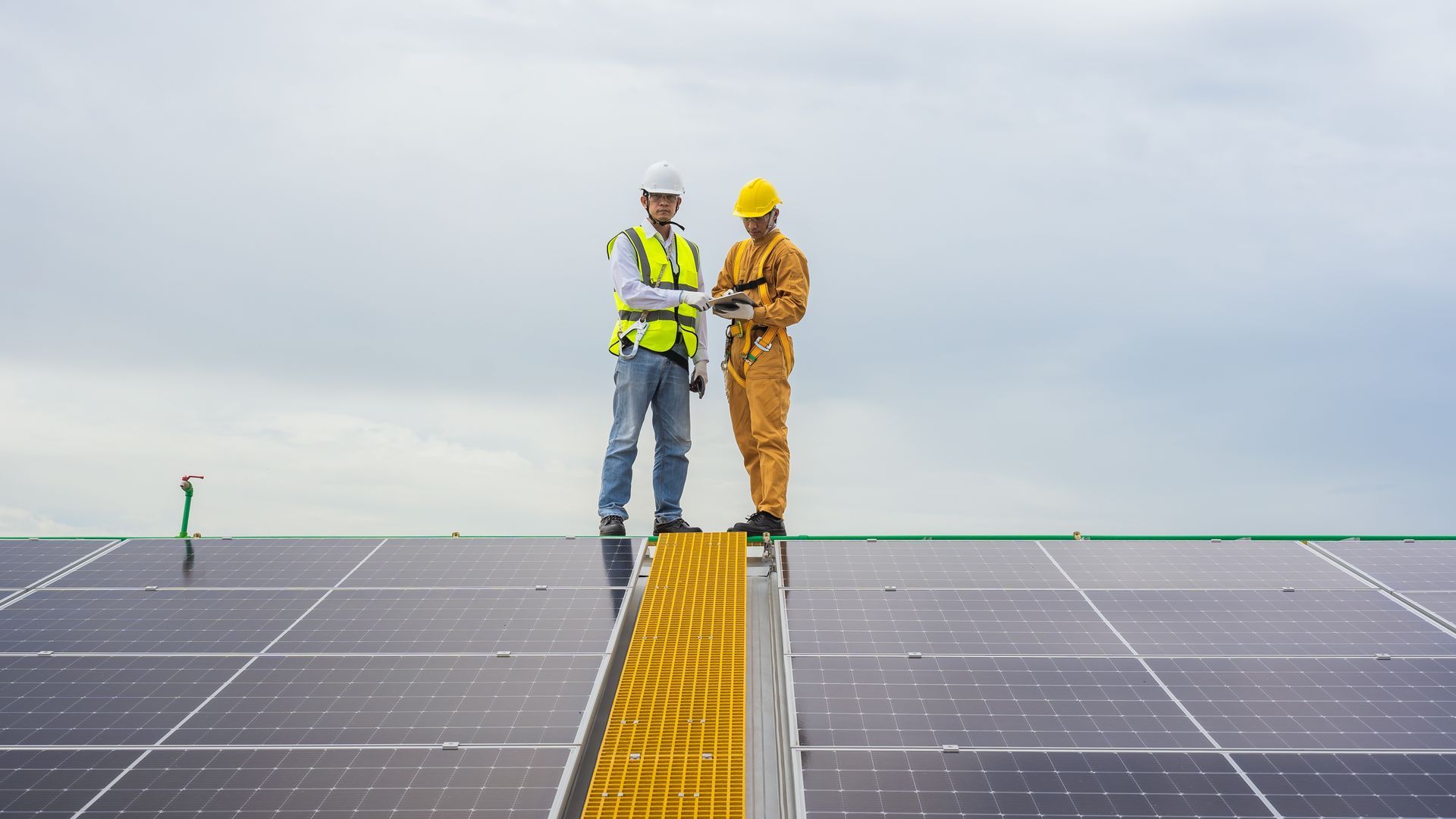 Two workers on solar panel roof, one pointing, other in safety gear, overcast sky.