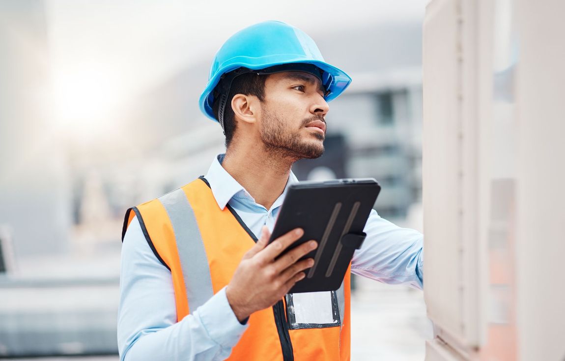 Construction worker in a blue helmet and orange vest, using a tablet, inspecting a structure outside.