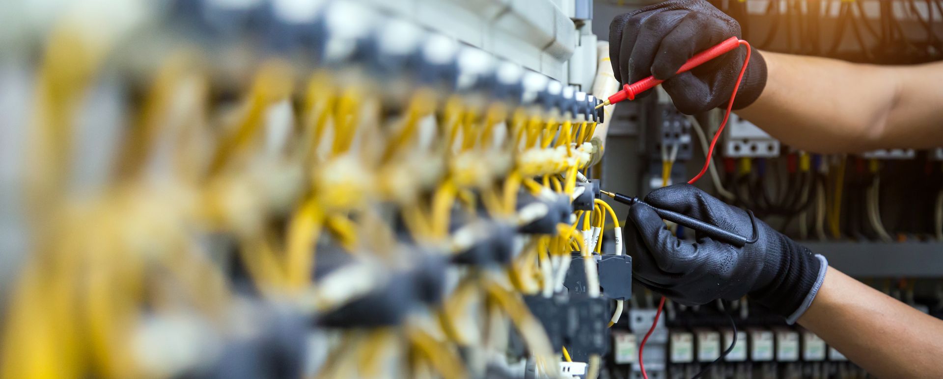 Hands wearing black gloves testing wires in a control panel with a multimeter.