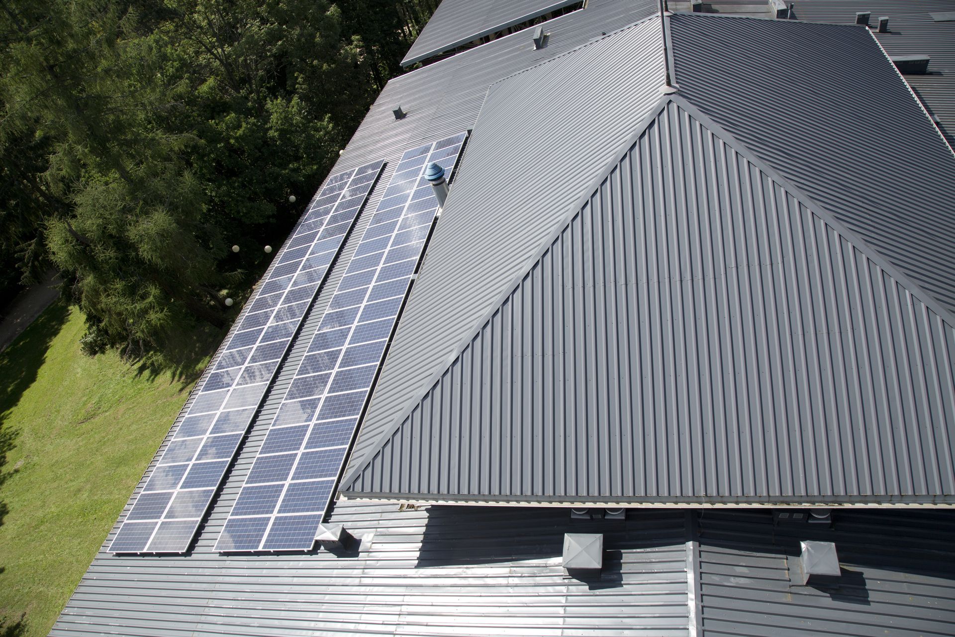 Solar panels on a gray metal roof of a building, next to a forested area.