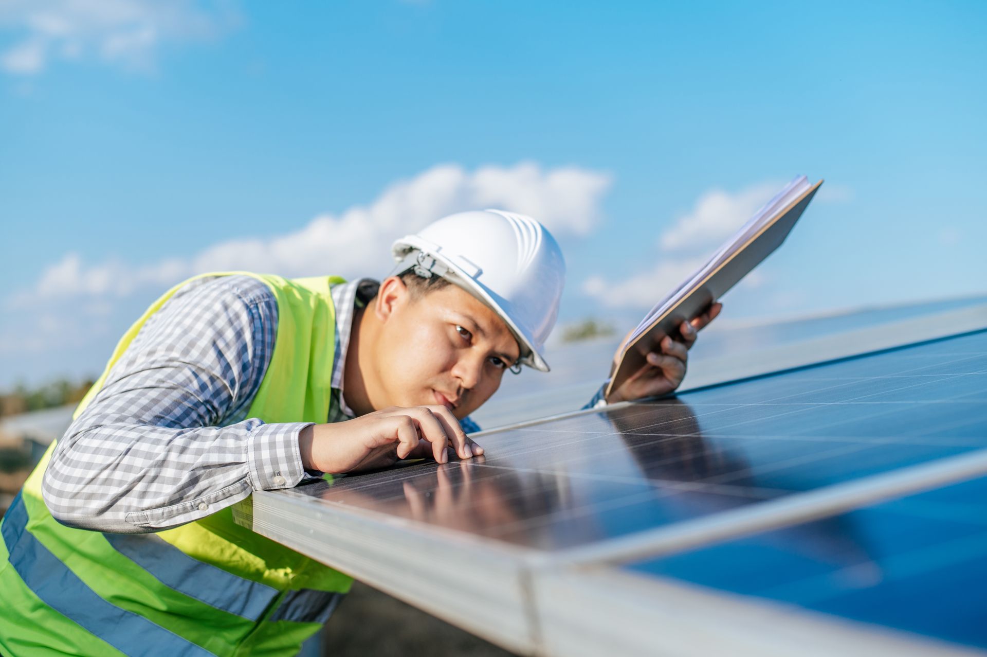 Technician in hard hat and safety vest inspecting solar panel, holding tablet outdoors under blue sky.