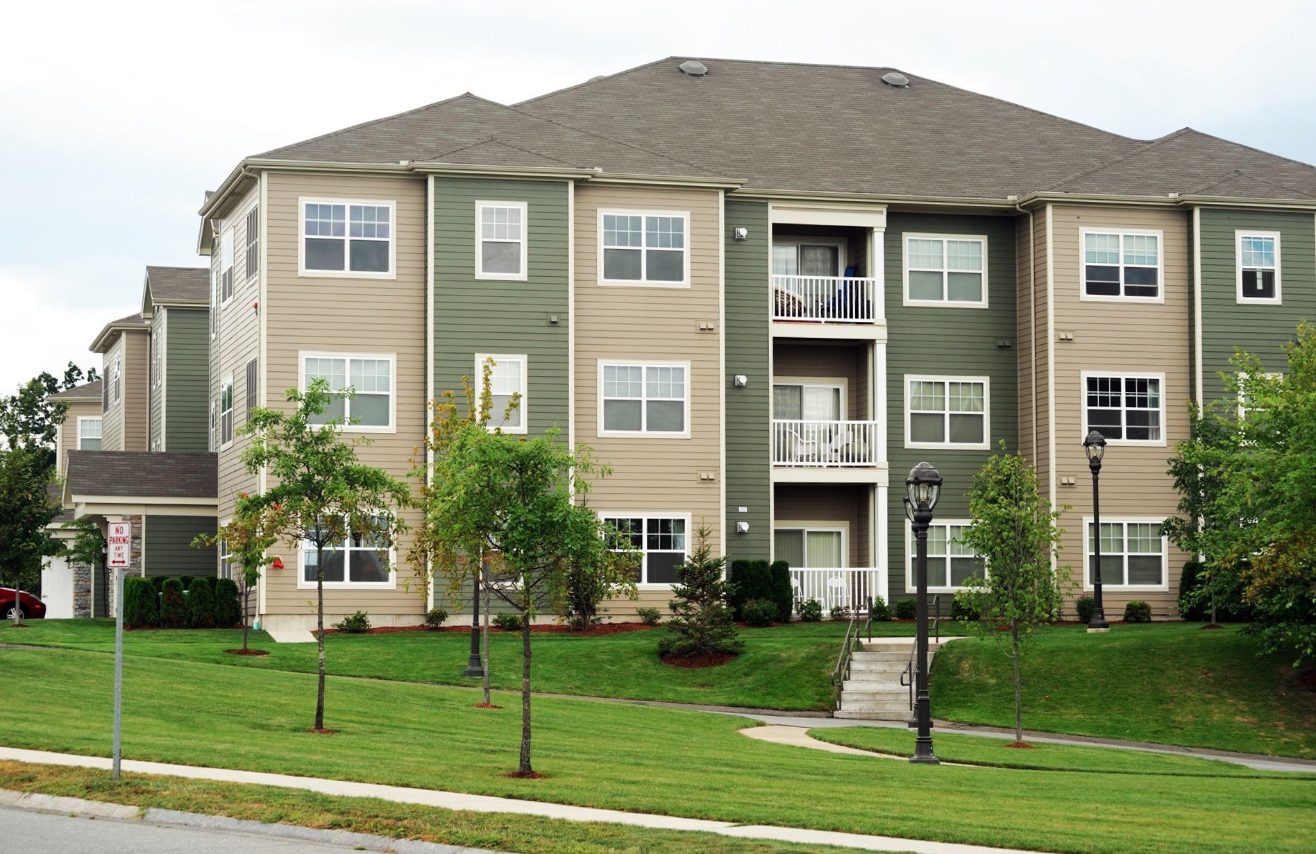 Three-story apartment building with green and tan siding, balconies, and a landscaped lawn in front