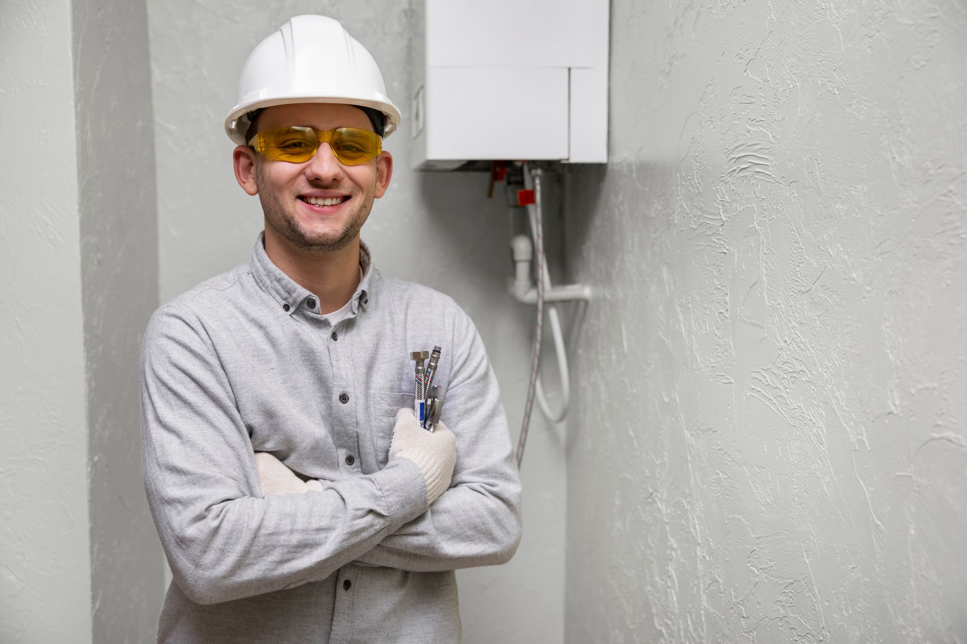Smiling technician in hard hat and safety glasses standing with arms crossed beside wall-mounted equipment