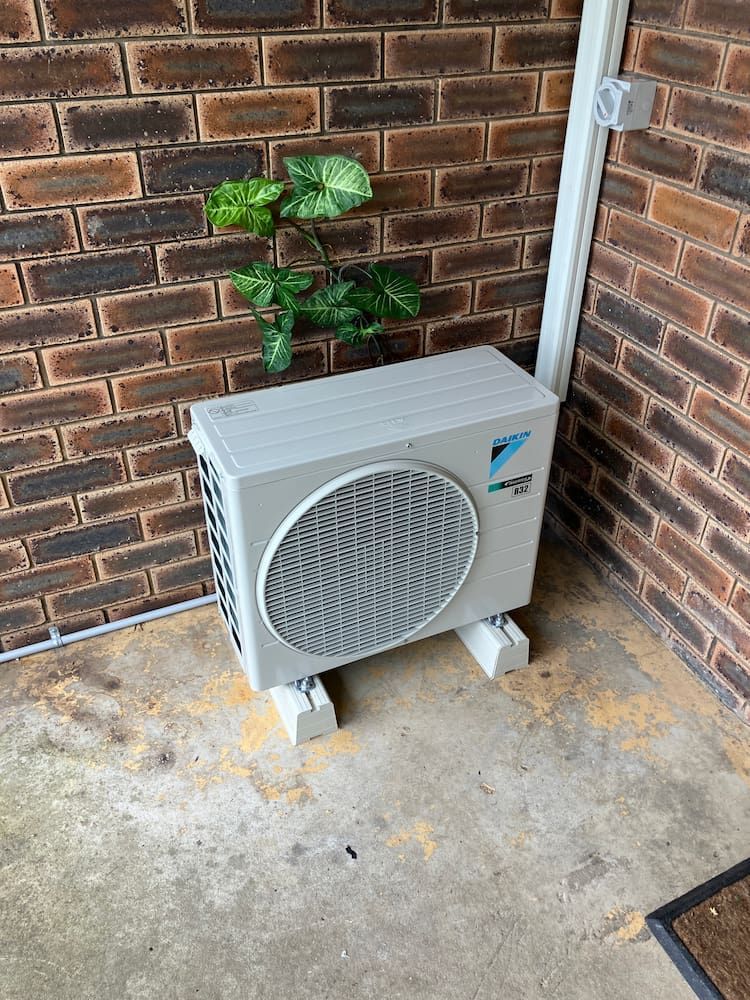 A White Air Conditioner is Sitting on the Ground Next to a Brick Wall — Comfort Solutions in Caloundra, QLD