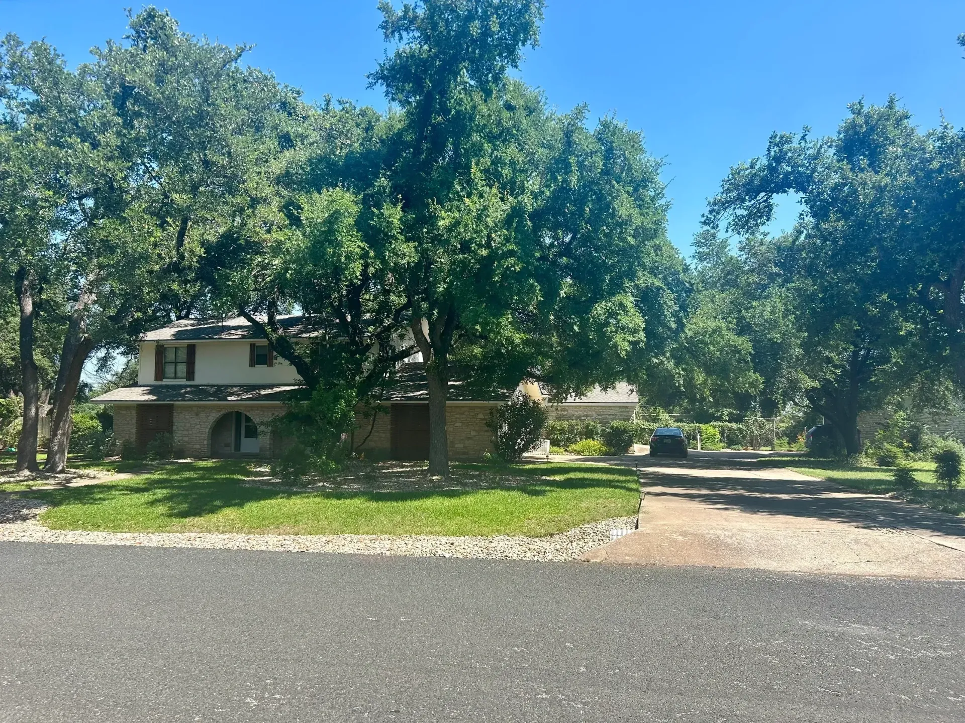 House with stone and light-colored siding, surrounded by large green trees, in bright daylight.
