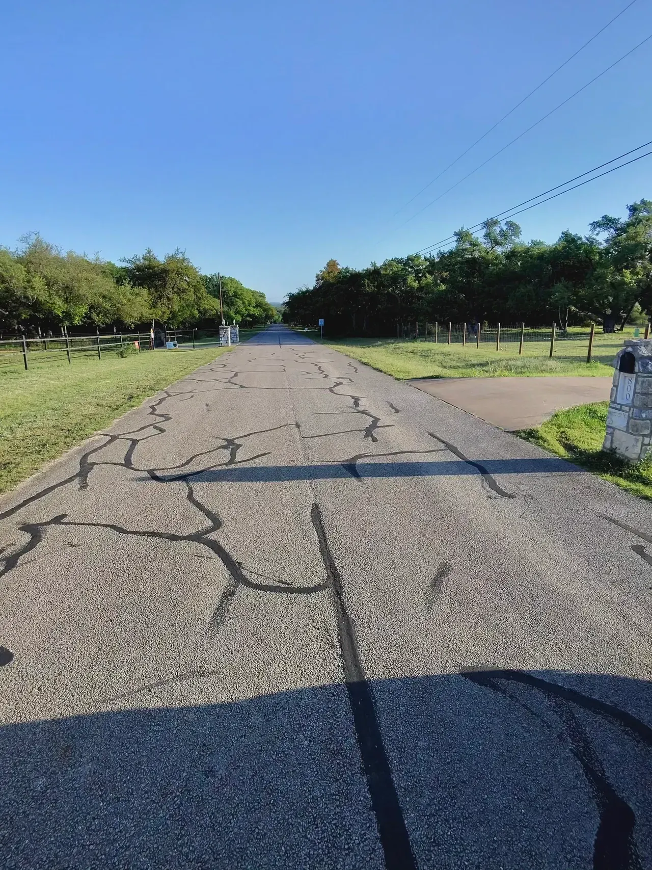 Cracked asphalt road leads to gate, surrounded by green trees and grass under a blue sky.