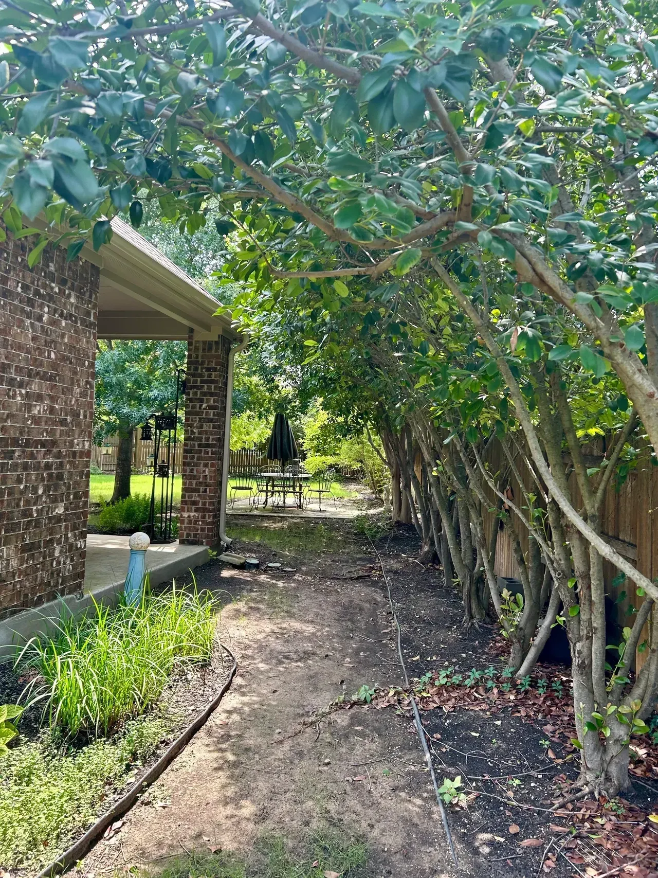 A dirt path leads past trees toward a brick building's shaded patio and a picnic table in a grassy yard.