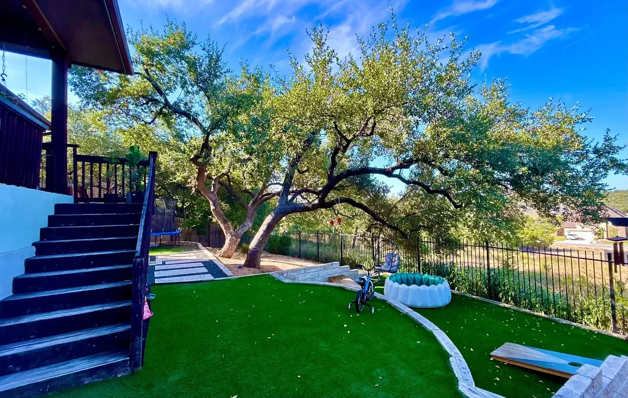 Backyard with a large tree, green lawn, stone walkway, and a deck with stairs under a blue sky.